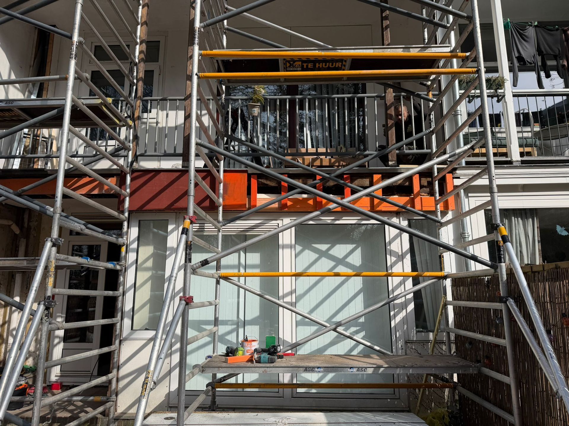 Steigers rondom een gebouwingang met oranje veiligheidshekken en bouwmaterialen in de buurt.