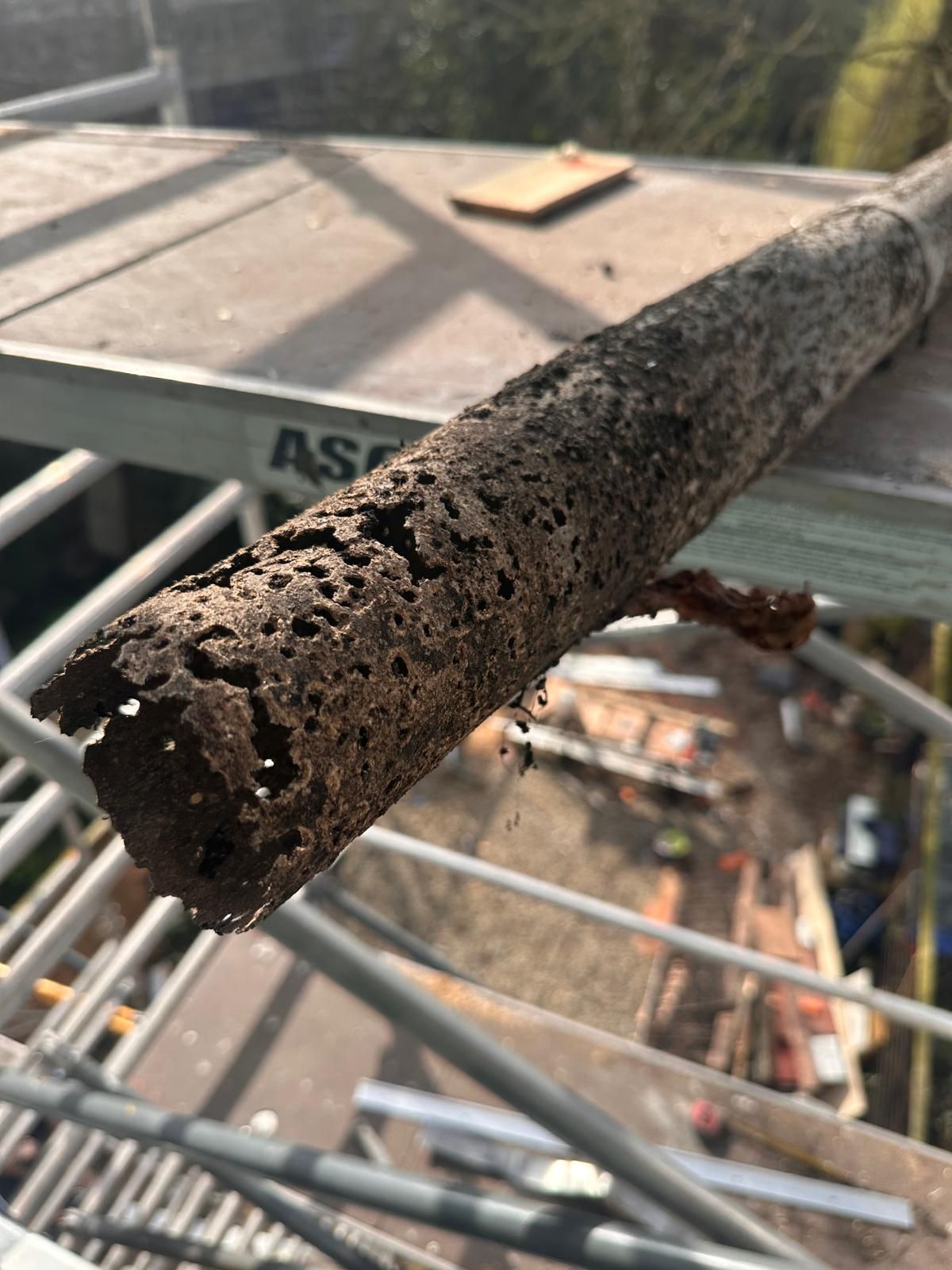 Een roestige metalen buis op een steiger, met op de achtergrond een dak en een bouwplaats.