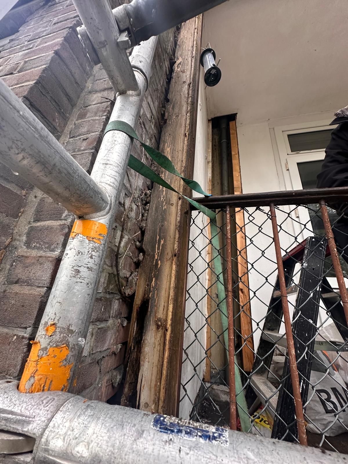 Smalle buitentrap met metalen steigers, gaashekwerk en een wandlamp boven een deuropening.