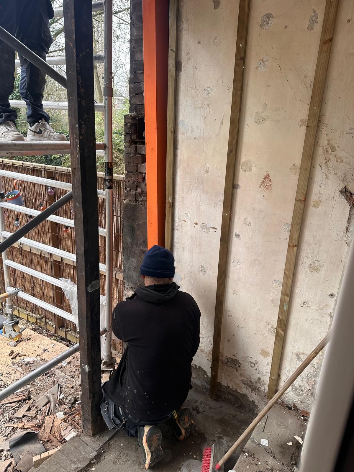 Een arbeider in een zwarte jas en blauwe muts hurkt naast een onafgewerkte muur op een bouwterrein.