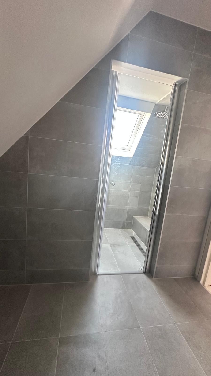 Kleine betegelde badkamer of doucheruimte met een gedeeltelijk openstaande glazen deur en een schuin plafond.