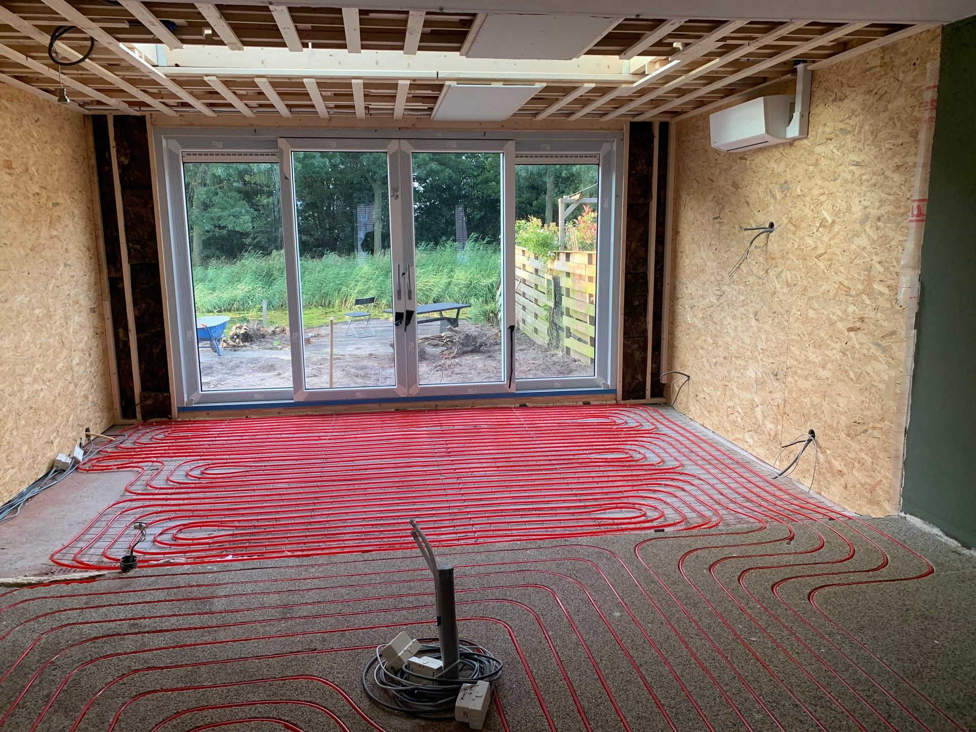 Renovatieruimte met rode vloerverwarmingsbuizen, glazen schuifdeuren en zichtbare plafondconstructie.