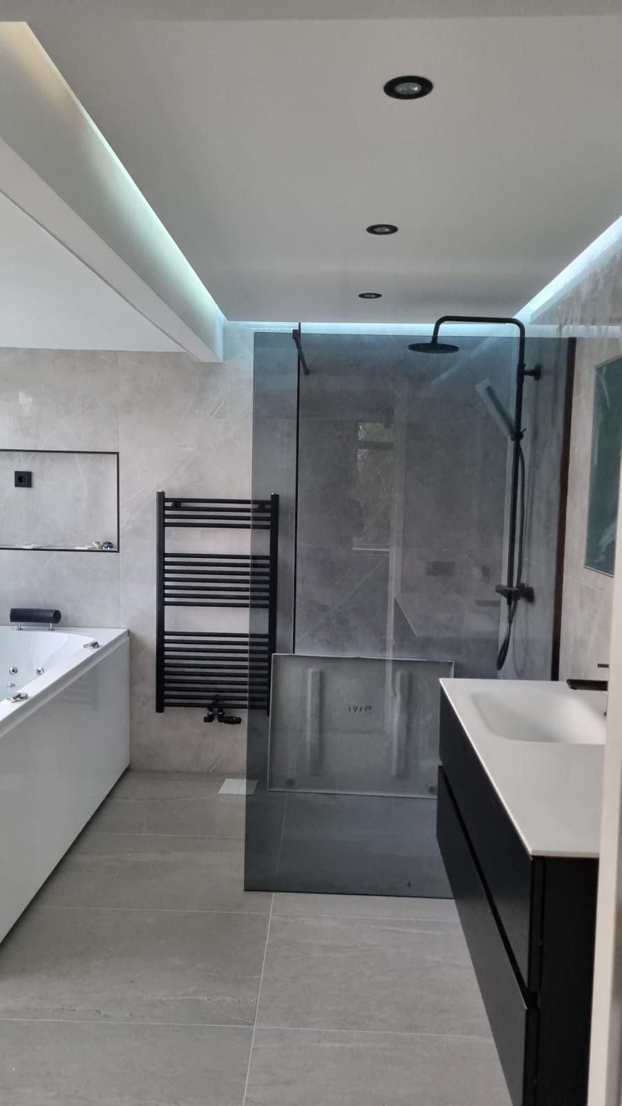 Moderne badkamer met glazen douchecabine, witte wastafel, badkuip en handdoekrek onder inbouwverlichting.