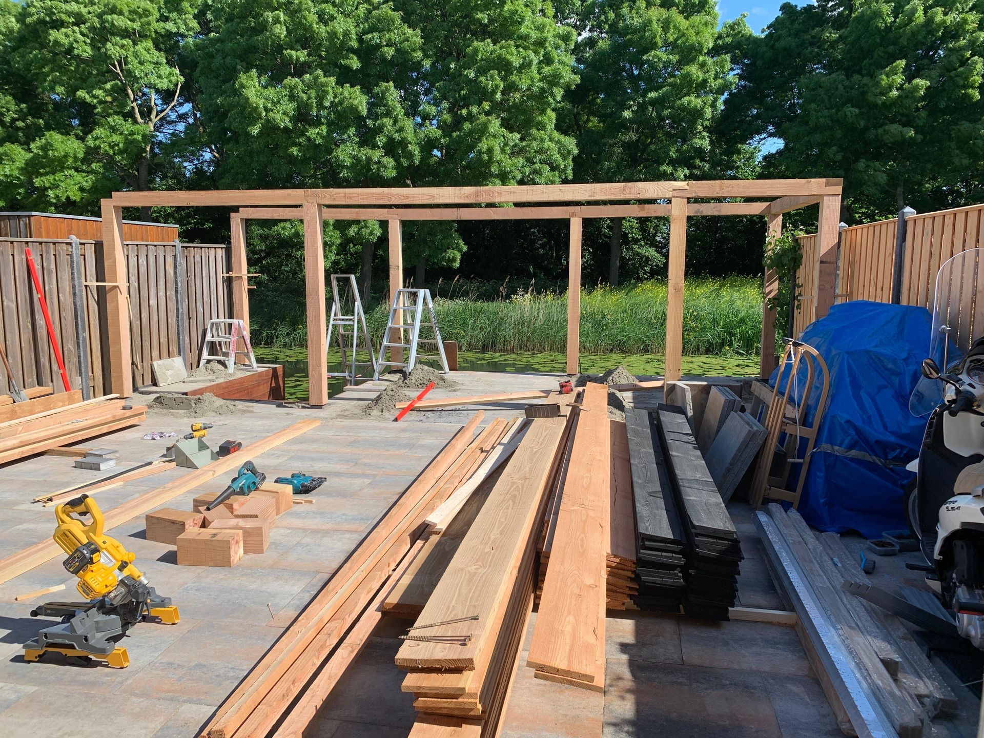 Een gedeeltelijk gebouwd houten terrasframe met hout, gereedschap en een ladder in een achtertuin in aanbouw.