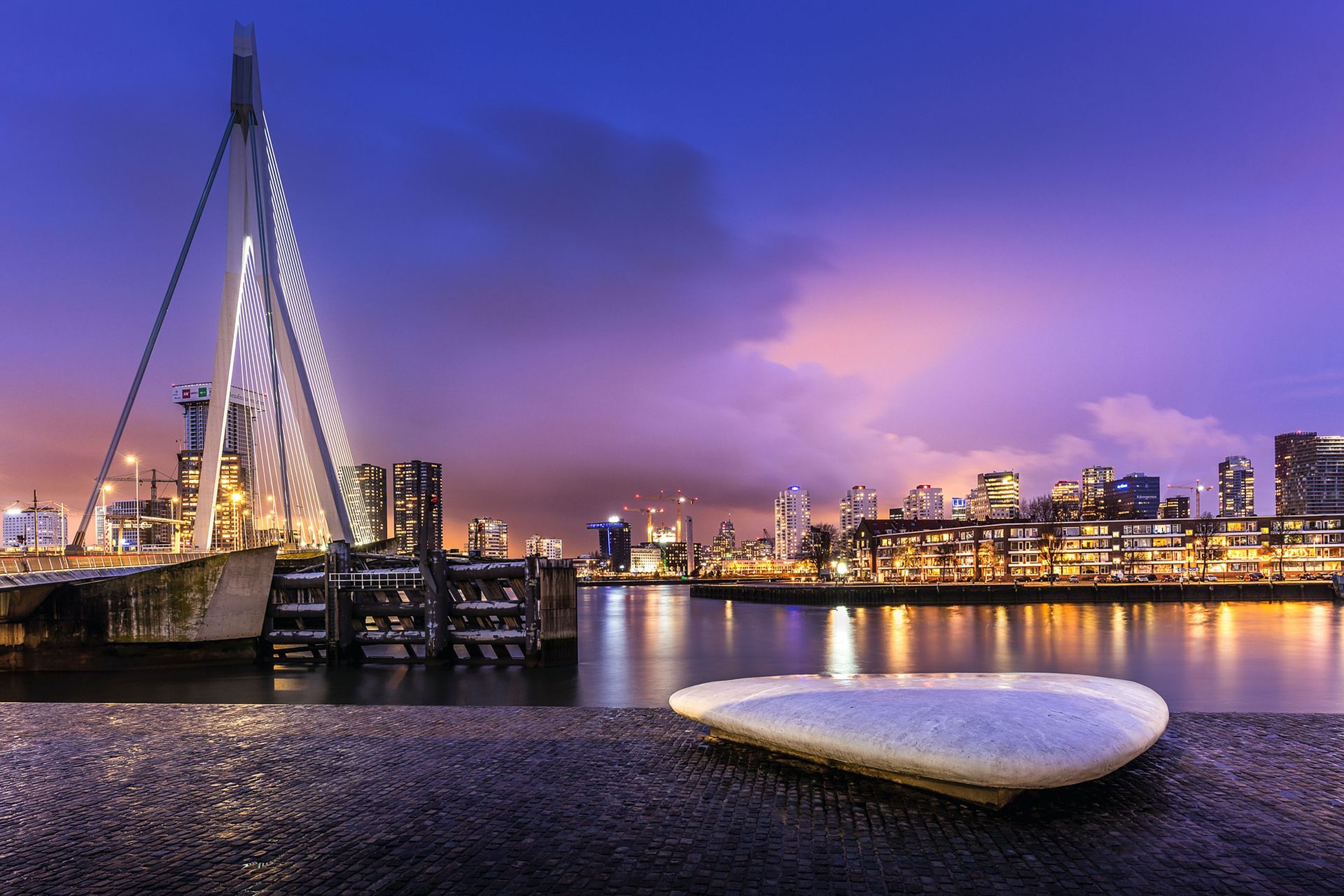 De skyline van de waterkant bij schemering, met brugverlichting en een witte boot op de voorgrond.