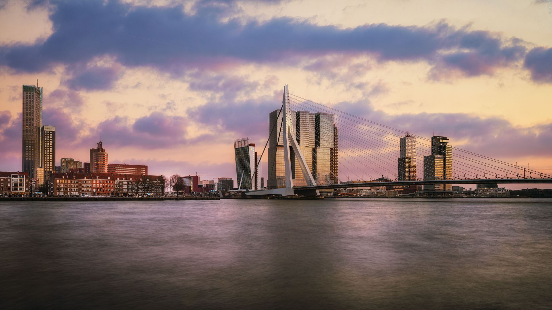 De skyline van de stad bij schemering, met een kalme waterkant, moderne torens en een brug onder roze en paarse wolken.
