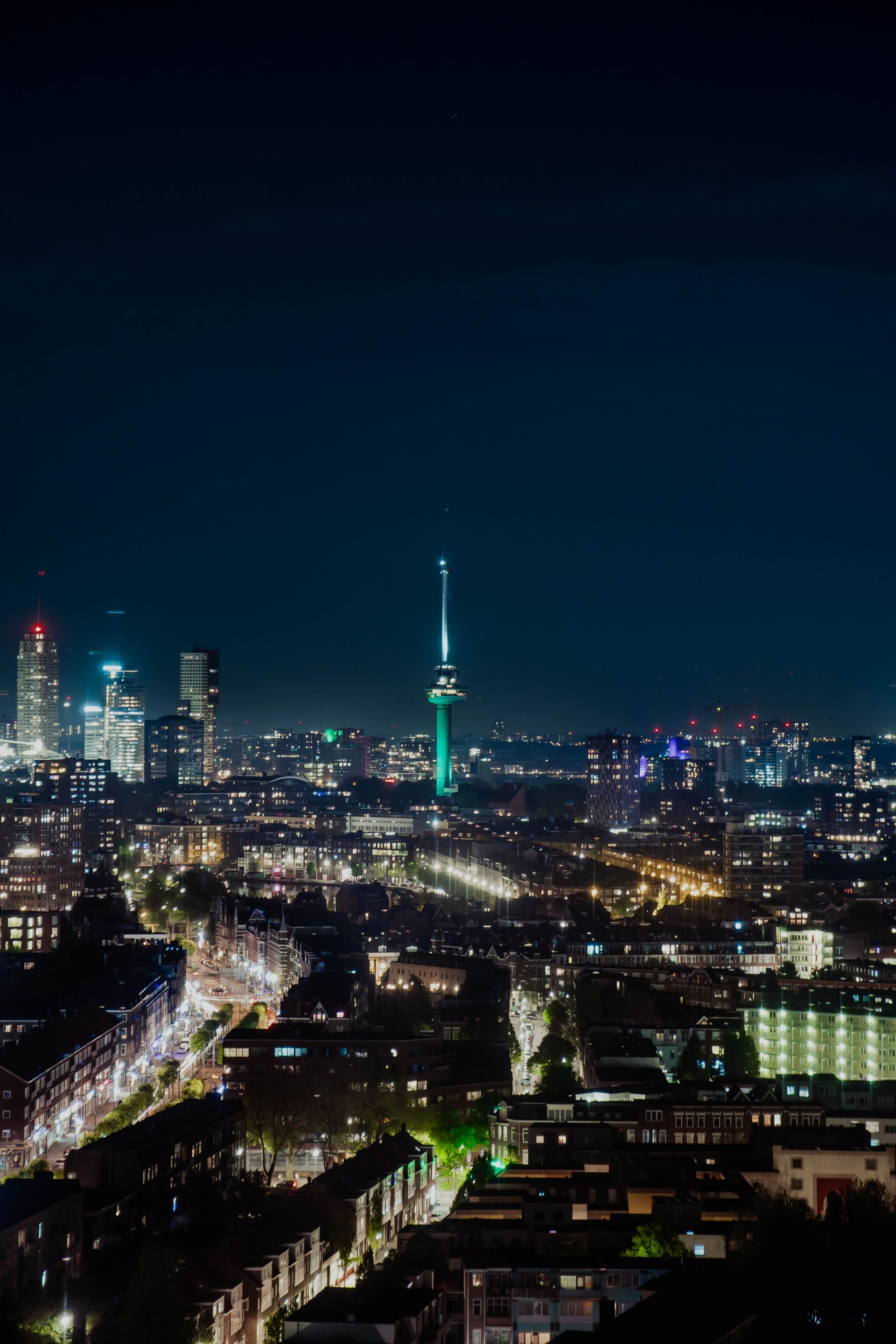 De nachtelijke skyline van de stad met een groen verlichte toren en gloeiende straten onder een donkere hemel.