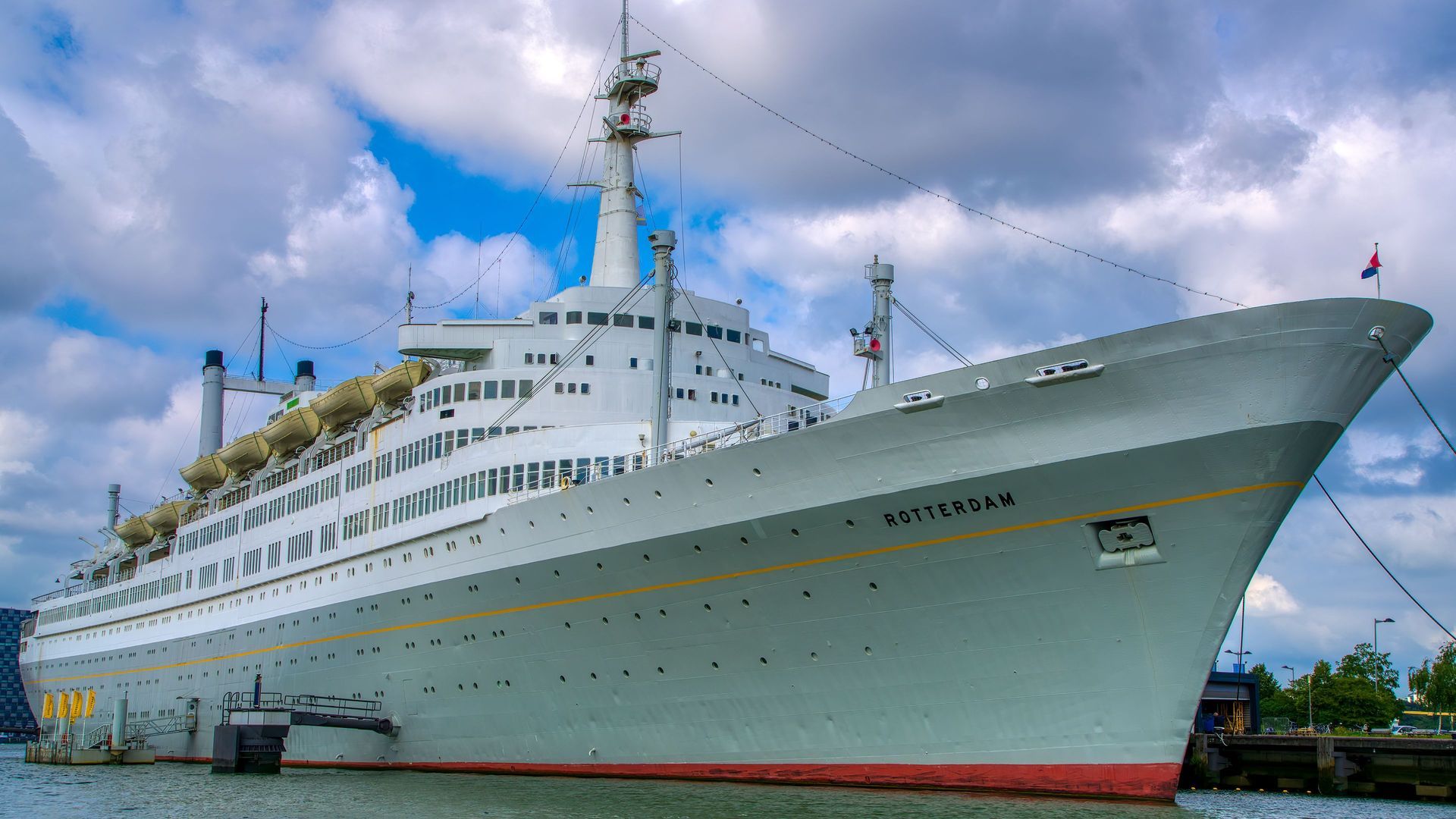 Een groot wit cruiseschip ligt aangemeerd in een haven onder een bewolkte blauwe hemel.