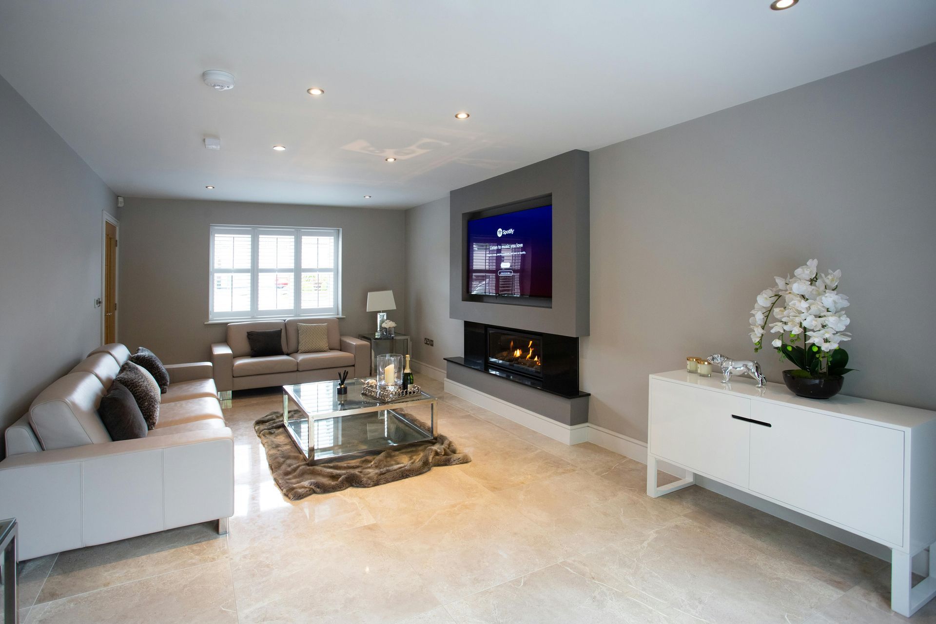 Moderne woonkamer met grijze bank, open haard, grote tv, salontafel en wit dressoir.