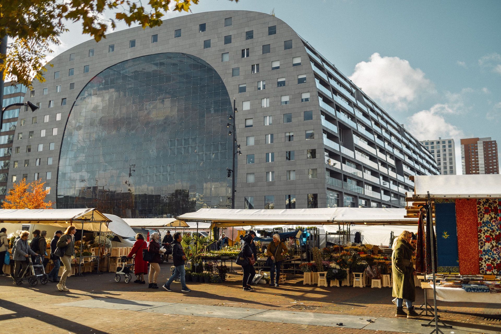 Modern gebouw van glas en staal achter een drukke openluchtmarkt met kraampjes en mensen onder witte tenten.