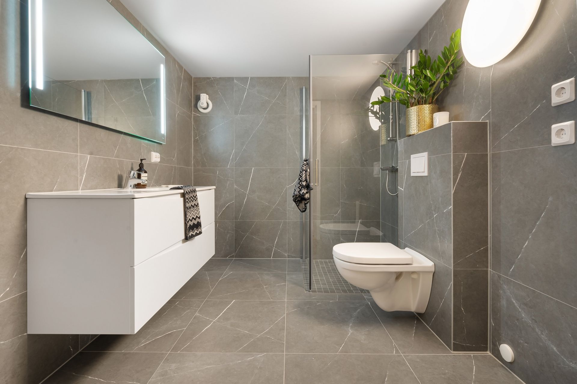Moderne badkamer met grijze tegels, zwevend wastafelmeubel, wandtoilet en glazen douchecabine.