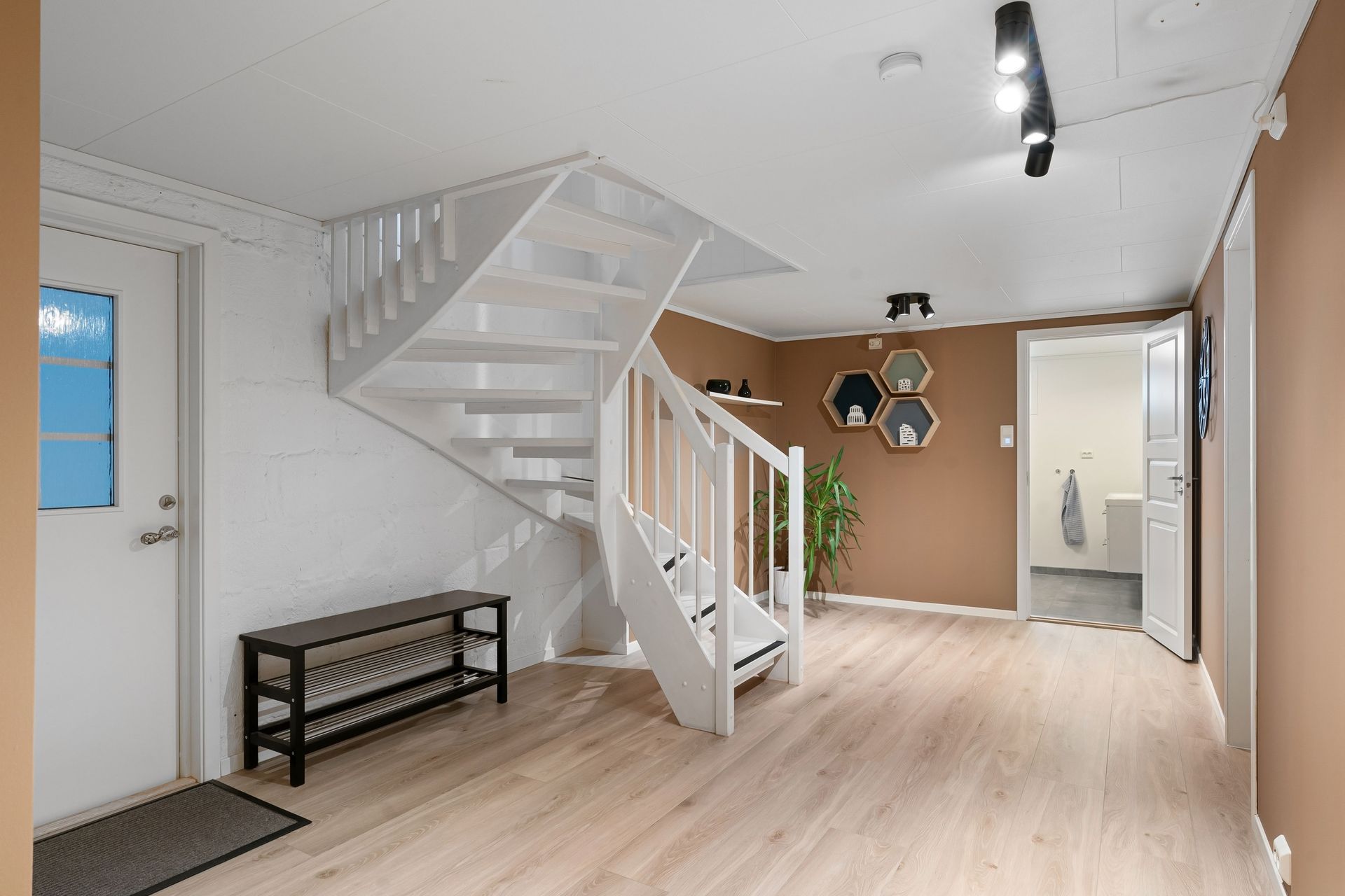 Lichte hal met witte trap, lichte houten vloer, bankje en muurdecoratie in een modern huis.