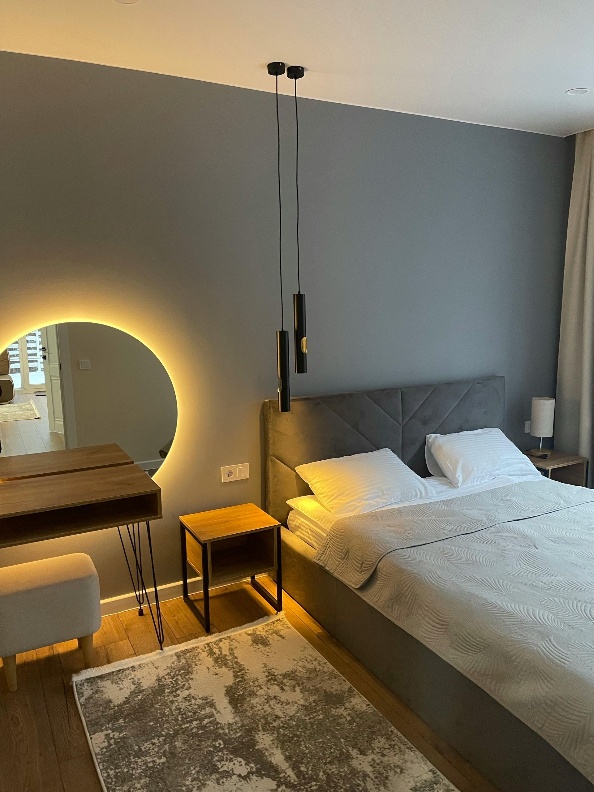 Moderne slaapkamer met grijs bed, glanzende ronde spiegel boven de wastafel, nachtkastje en staande lamp op een vloerkleed.