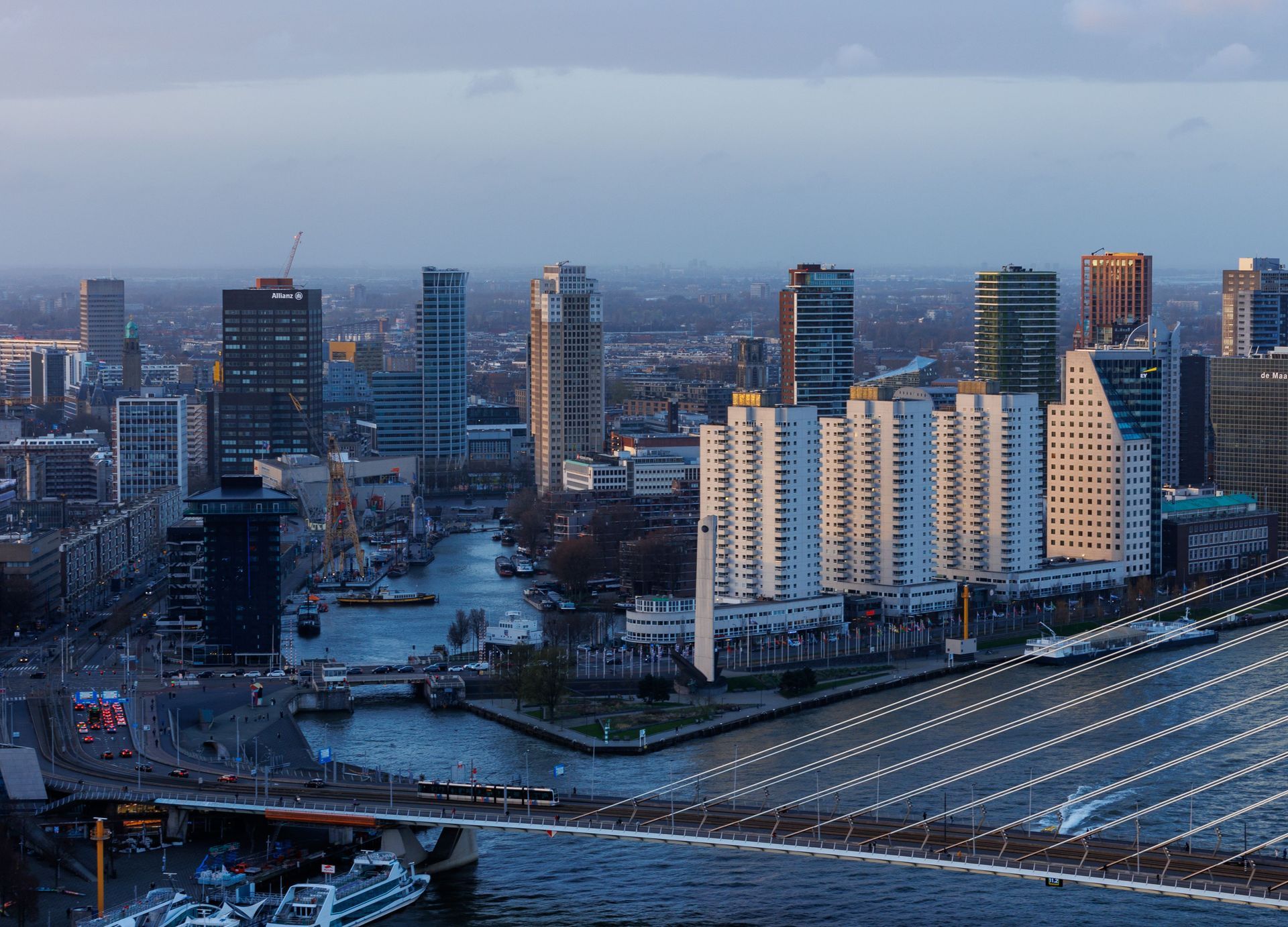 Stadsgezicht bij schemering met flatgebouwen, een rivier en een verlichte brug op de voorgrond.