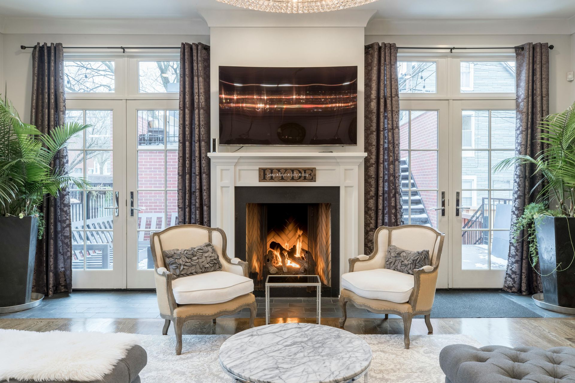 Elegante woonkamer met een brandende open haard, een aan de muur gemonteerde tv en twee fauteuils bij hoge ramen.