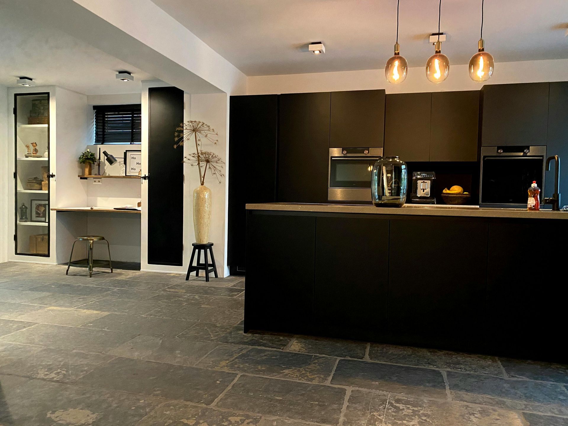 Moderne keuken met zwart kookeiland, hanglampen en een kleine ingebouwde bureauhoek aan de linkerkant.