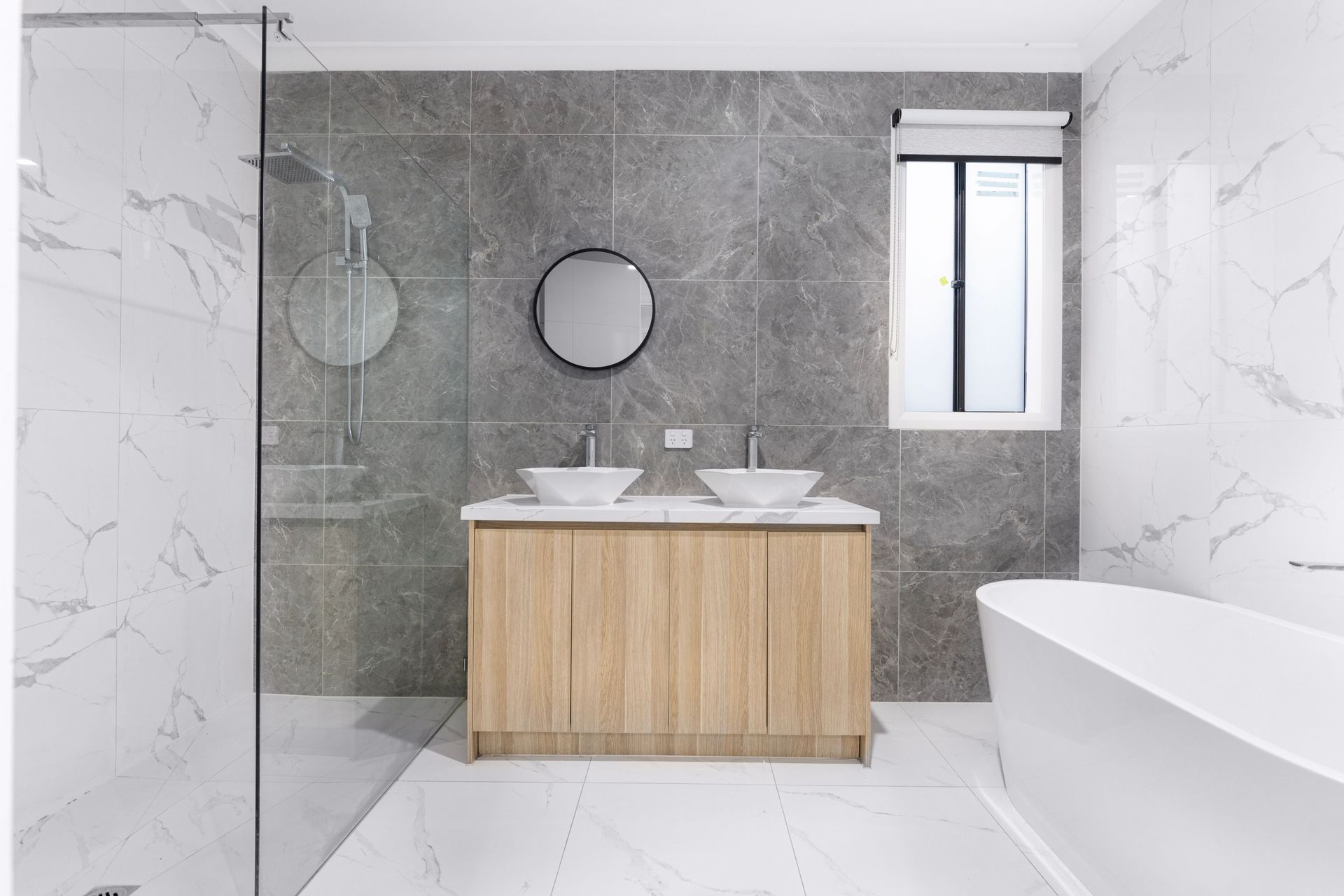 Moderne badkamer met dubbele wastafel, grijze tegelwanden, glazen douchecabine en vrijstaand bad.