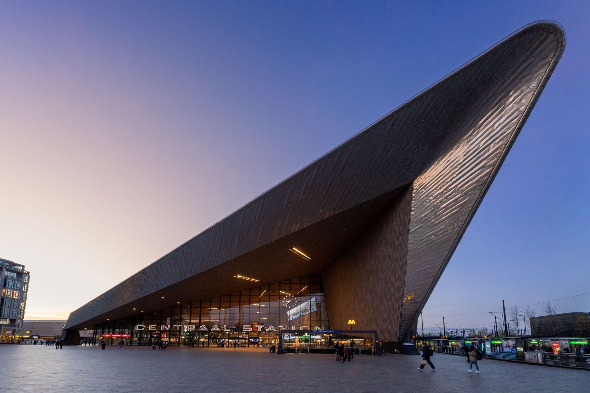 Modern, hoekig gebouw in de schemering met een schuin dak en verlichte ramen op een openbaar plein