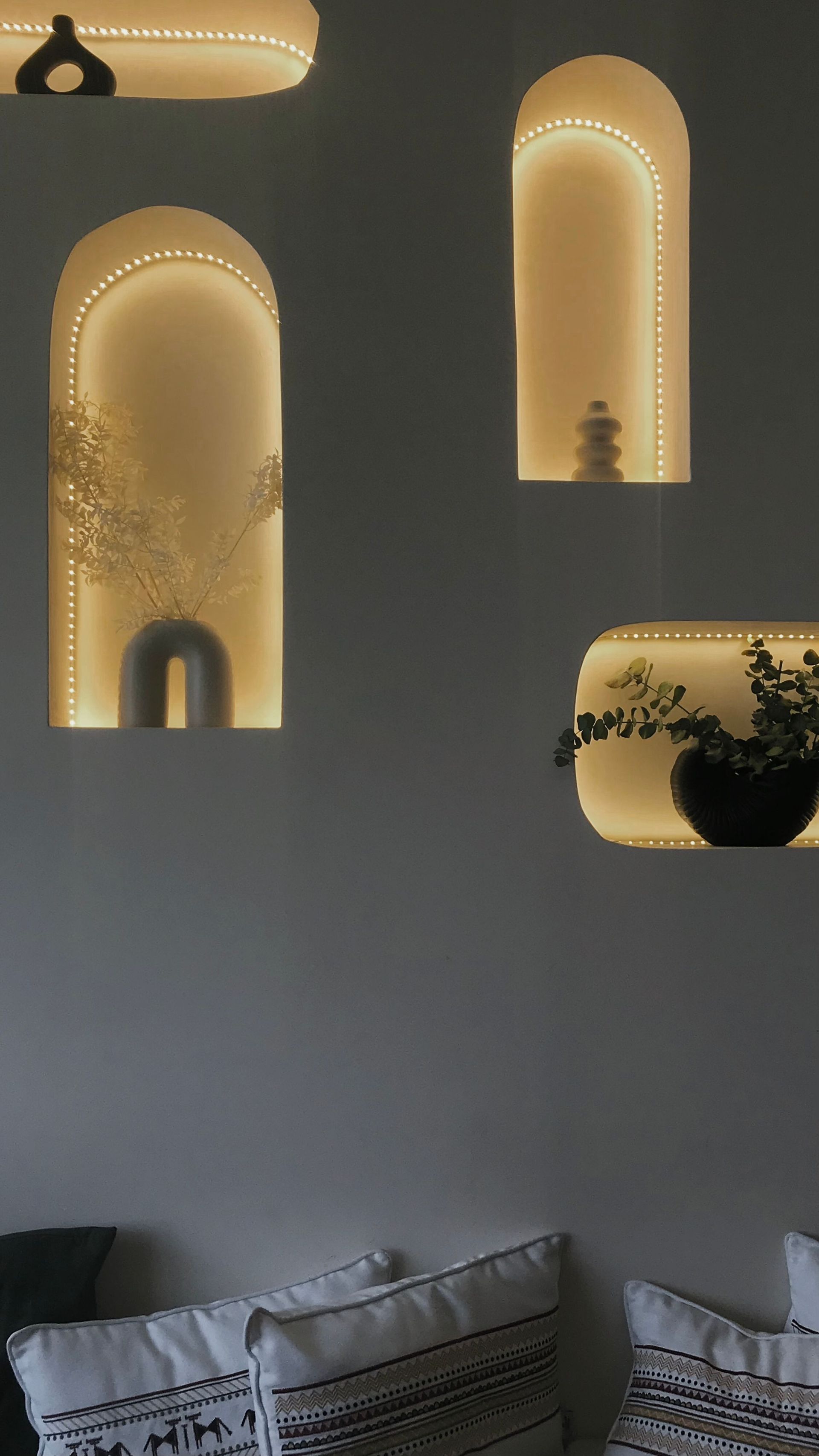 Moderne wand met warme, gebogen nisverlichting, planten en kussens eronder.