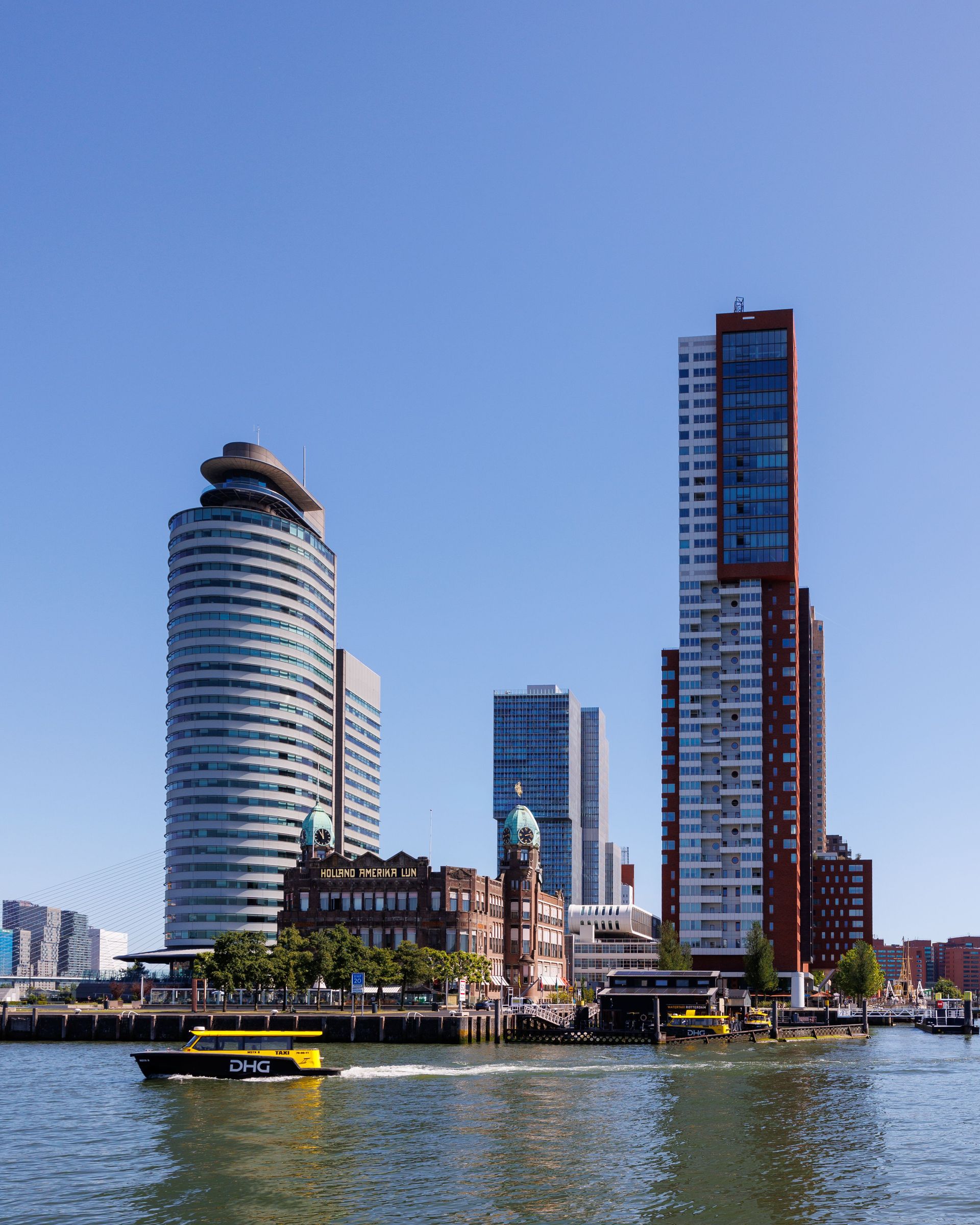Moderne skyline aan het water met hoge torens en een gele boot op het water onder een helderblauwe hemel.