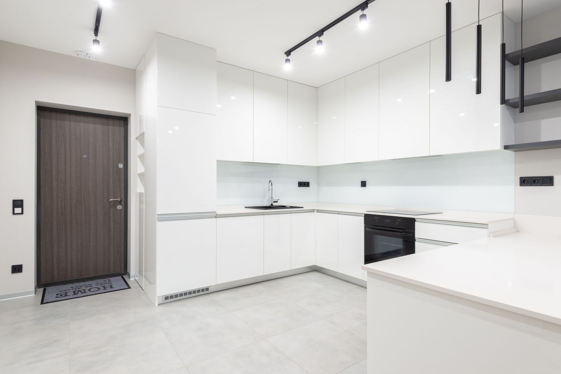 Moderne witte keuken met glanzende kasten, ingebouwde oven, zwarte railverlichting en een houten voordeur.