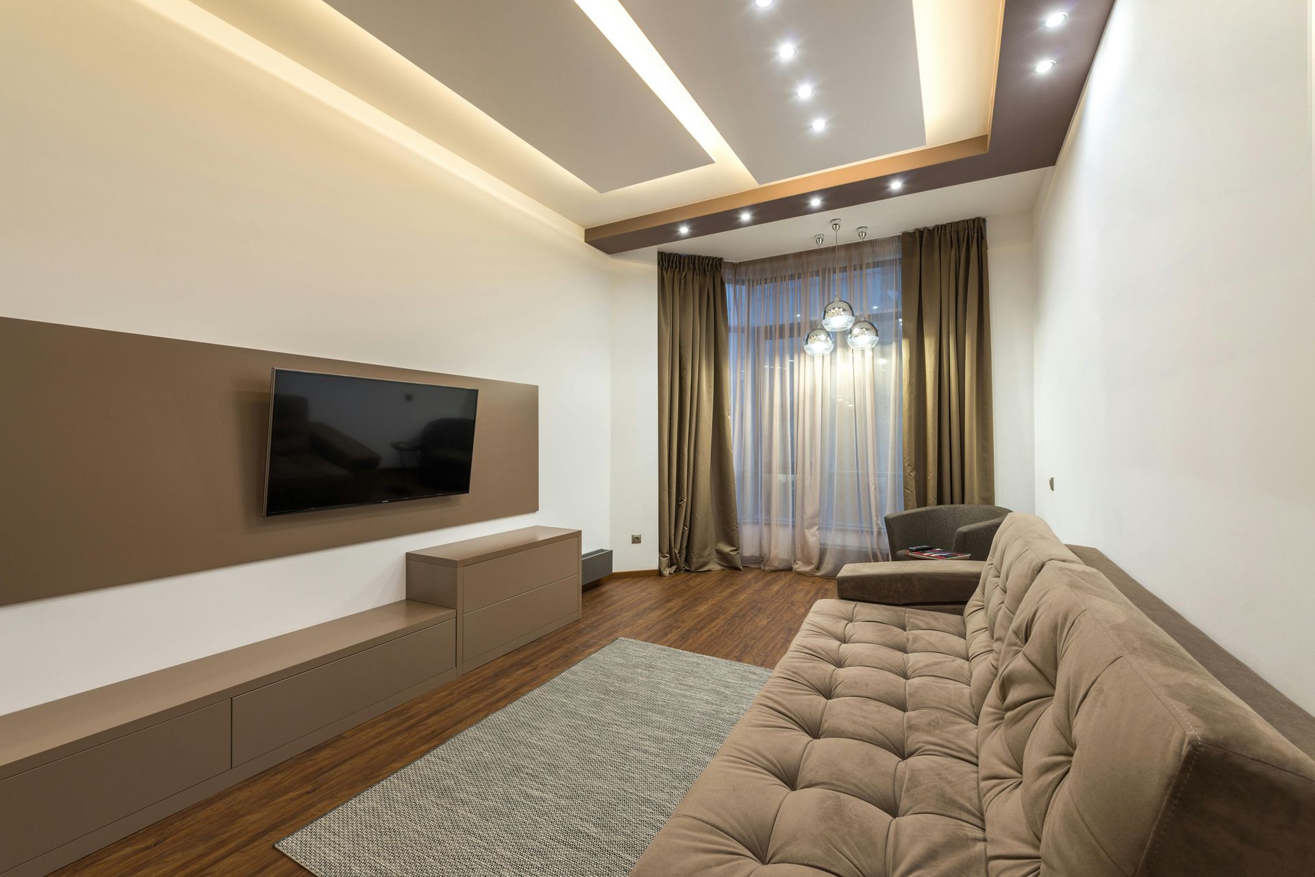 Moderne woonkamer met beige bank, aan de muur gemonteerde tv, vloerkleed met patroon en zachte plafondverlichting.