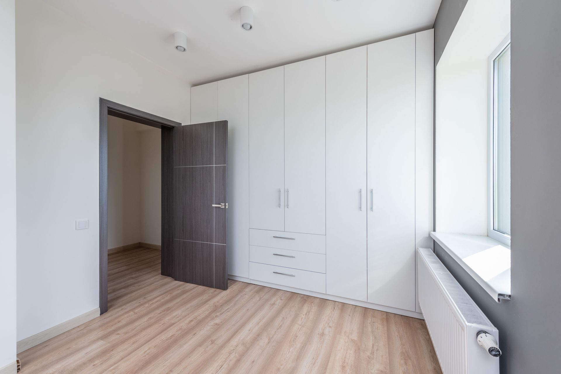 Lege witte slaapkamer met houten vloer, ingebouwde kast, open deur en raamradiator boven de vensterbank.