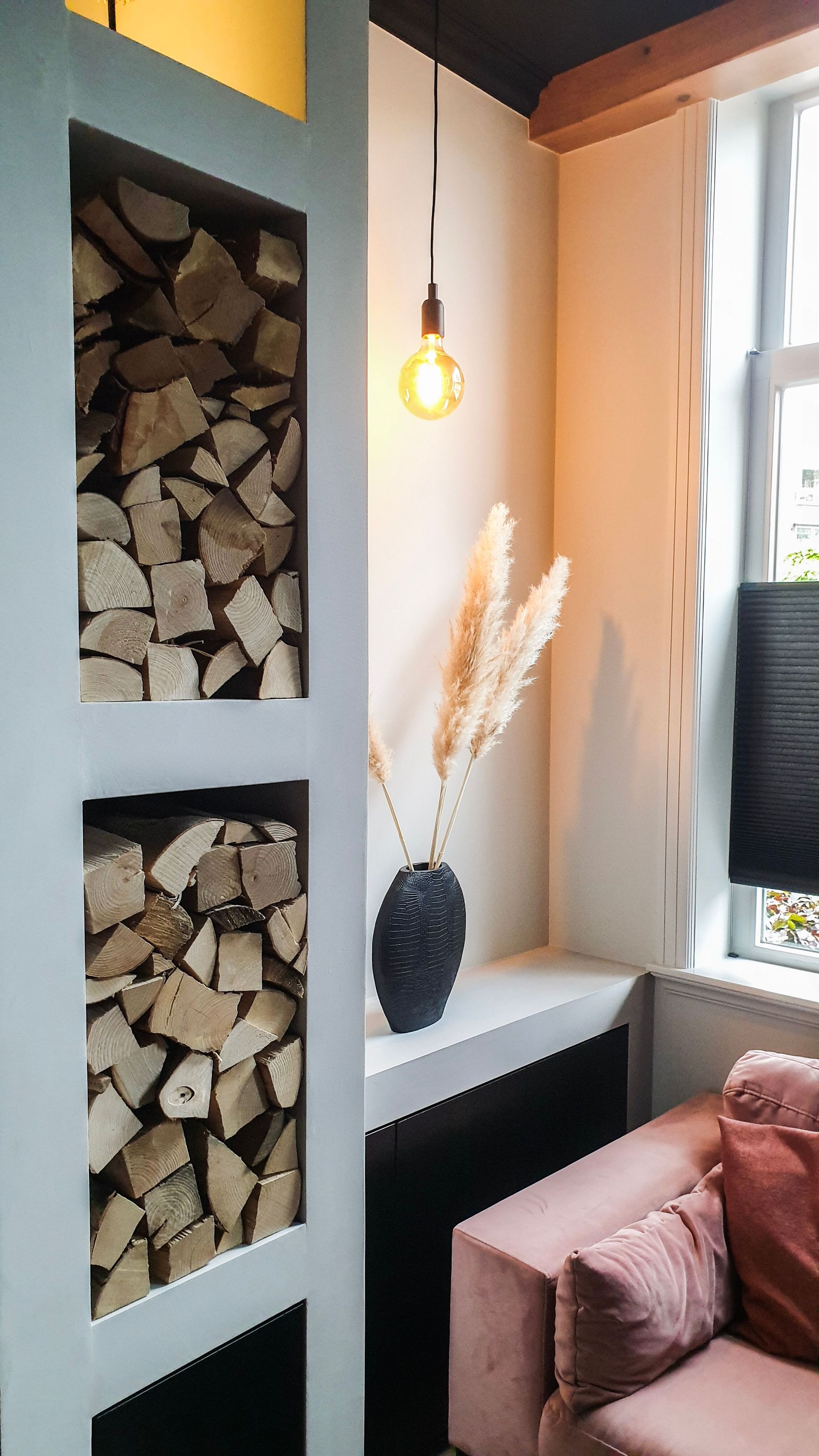 Gezellig hoekje met planken vol brandhout, een hanglamp, een witte vaas met pampasgras en een roze bank bij het raam.