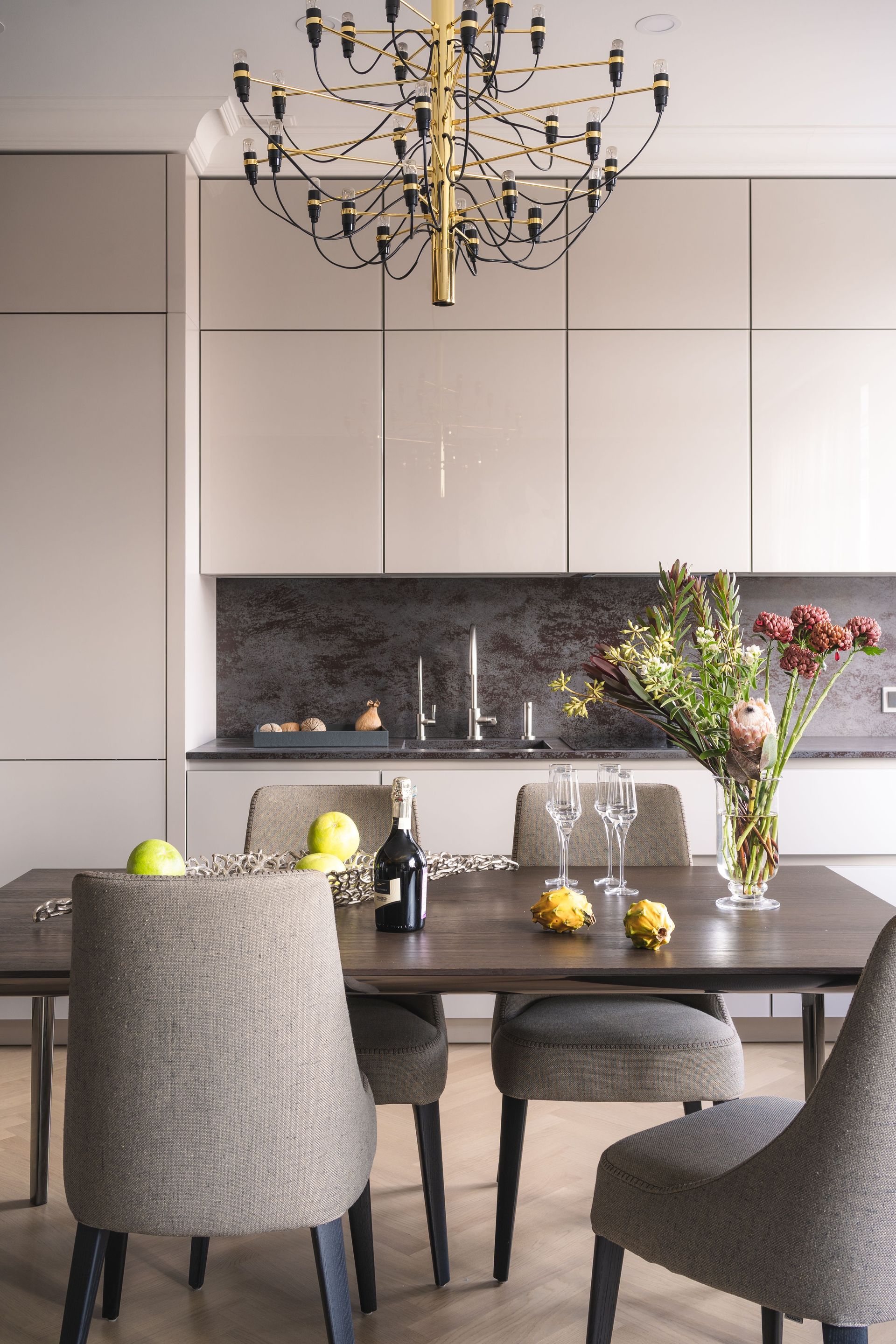 Moderne eetkamer met een donkere tafel, grijze stoelen, bloemen, fruit en een kroonluchter.