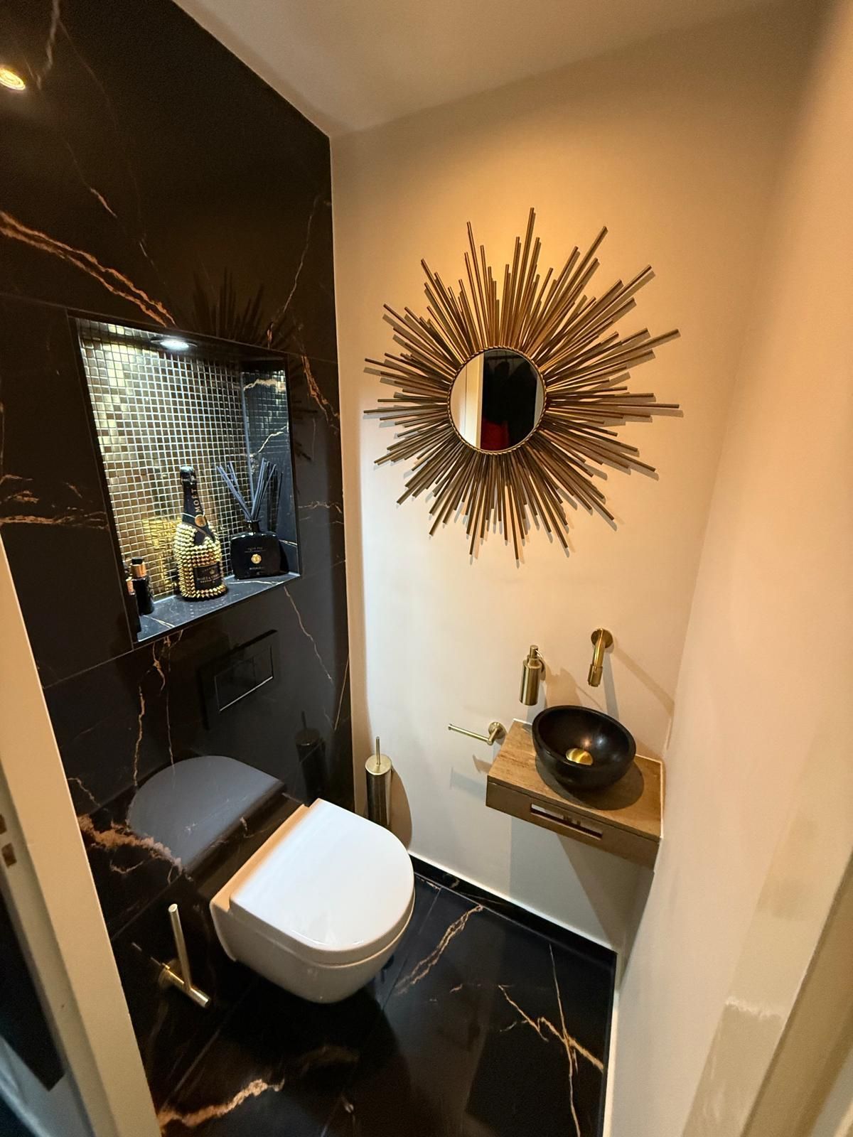 Moderne zwart-gouden badkamer met wandtoilet, ronde spiegel en wastafel op een houten badmeubel.