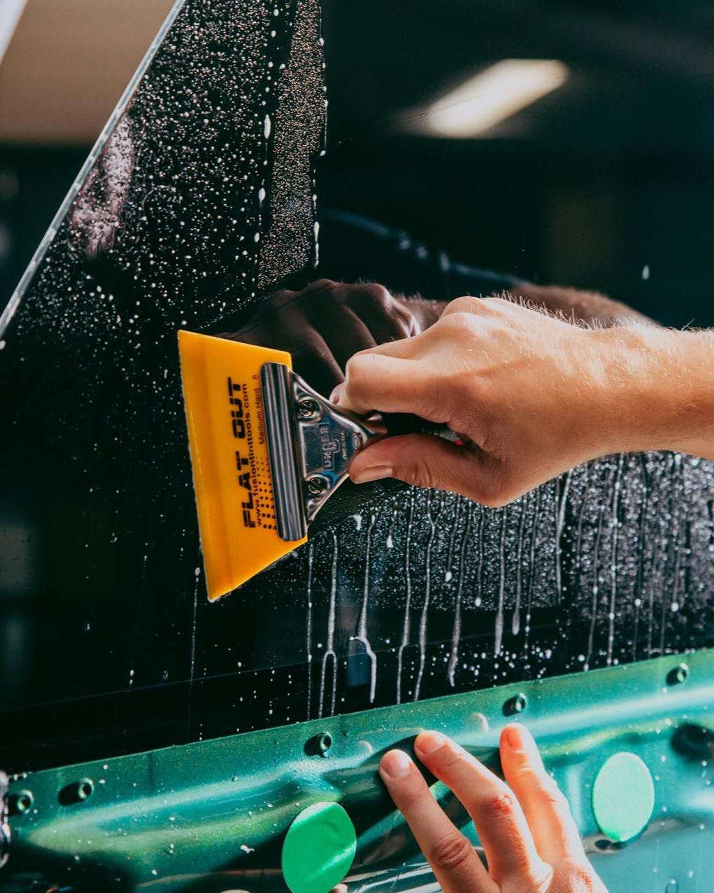 Hands using a yellow squeegee to clean a wet car window with water droplets.