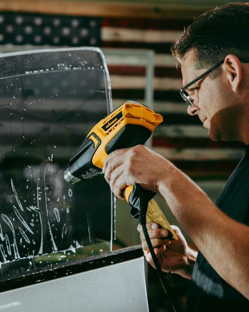 Man using a yellow heat gun to remove adhesive from a glass panel in a workshop