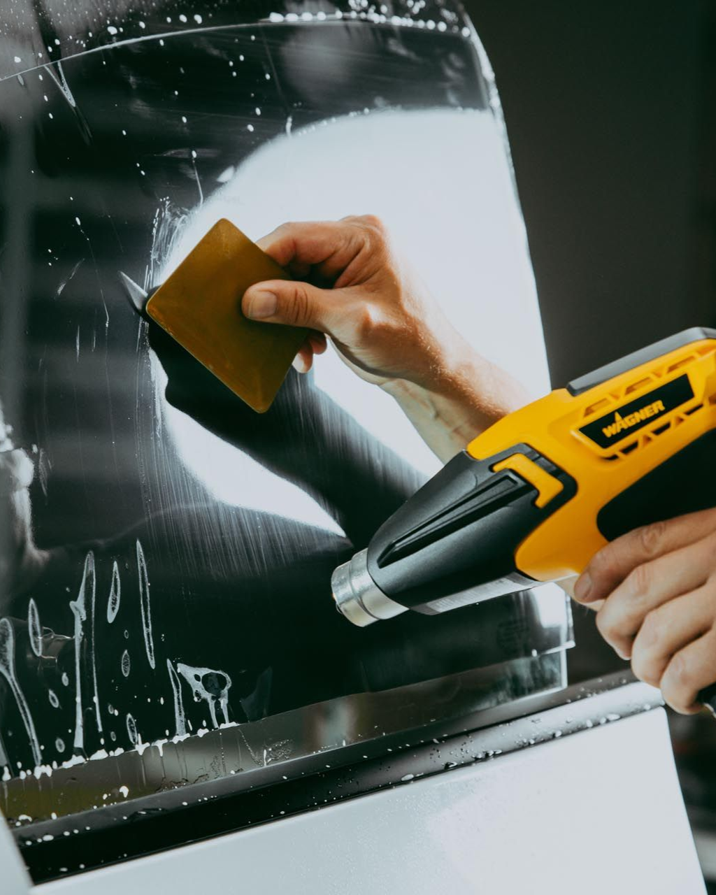 Hand using a scraper and heat gun to remove film from a glass surface