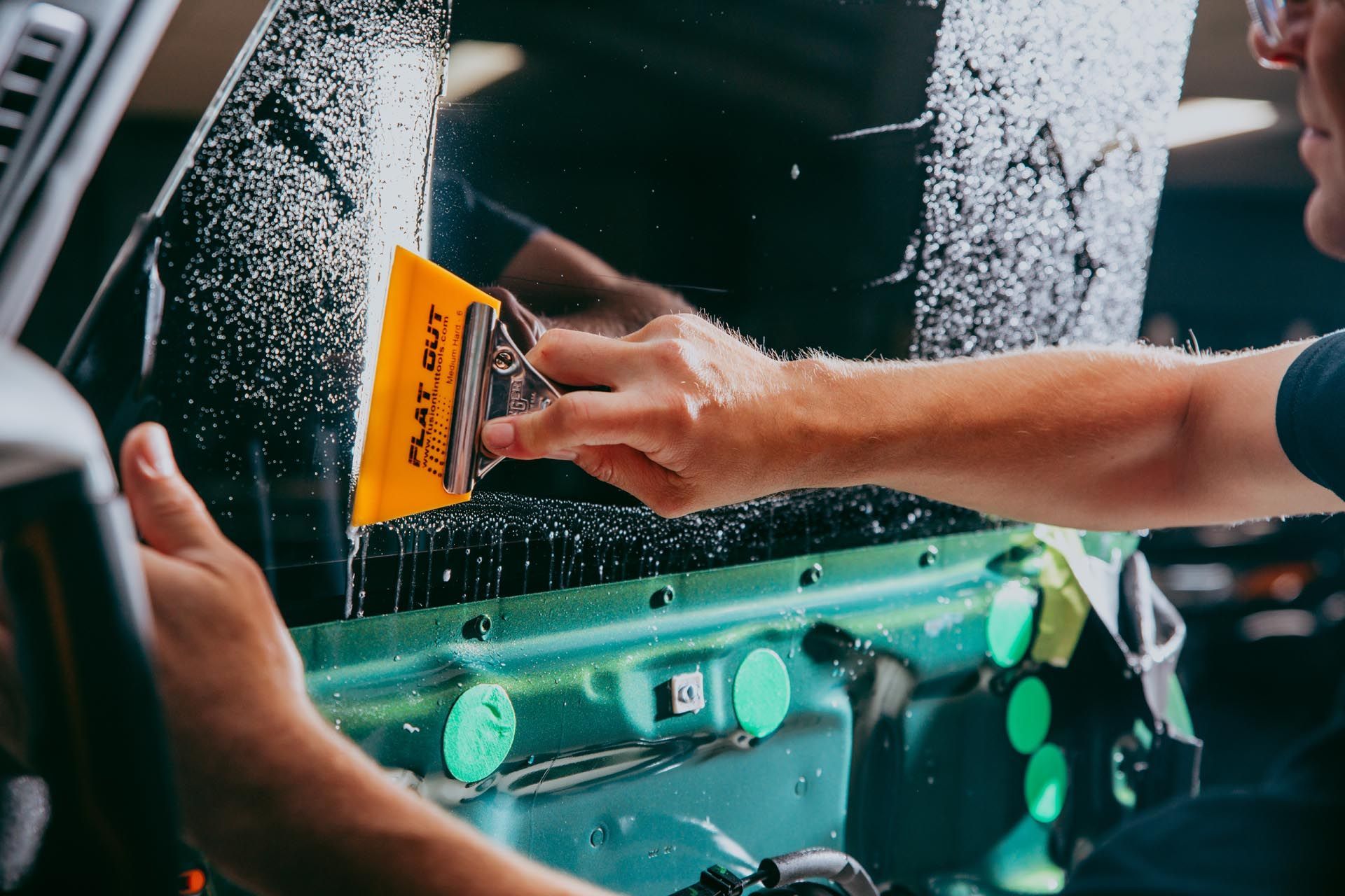 Hands applying a yellow squeegee to a car window, smoothing water and bubbles during tint or vinyl installation