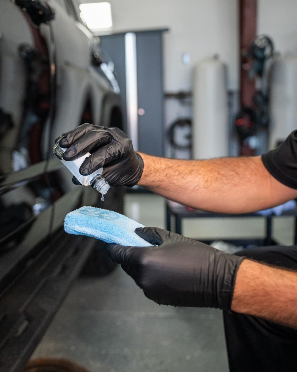 Gloved hands cleaning a car’s black side panel with a blue sponge in a garage