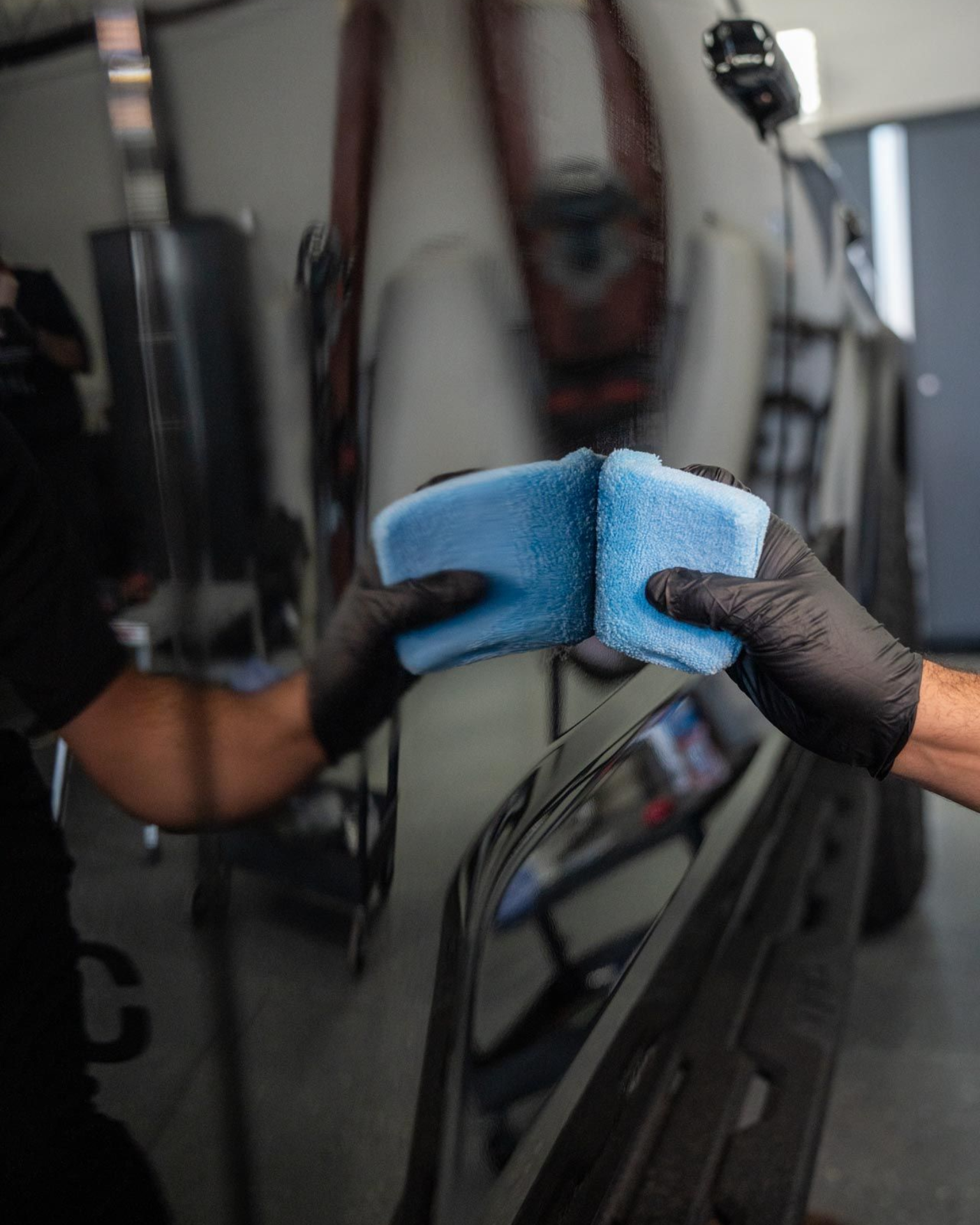 Gloved hands holding a blue microfiber cloth against a car windshield in a garage