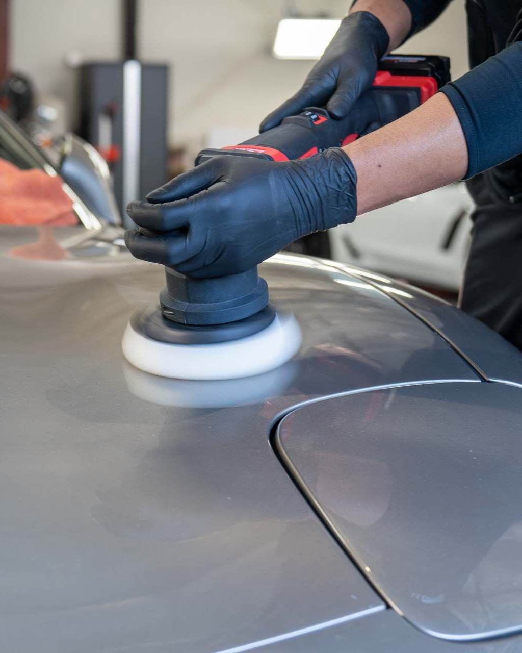 Gloved hands polishing a gray car with a buffer in a garage