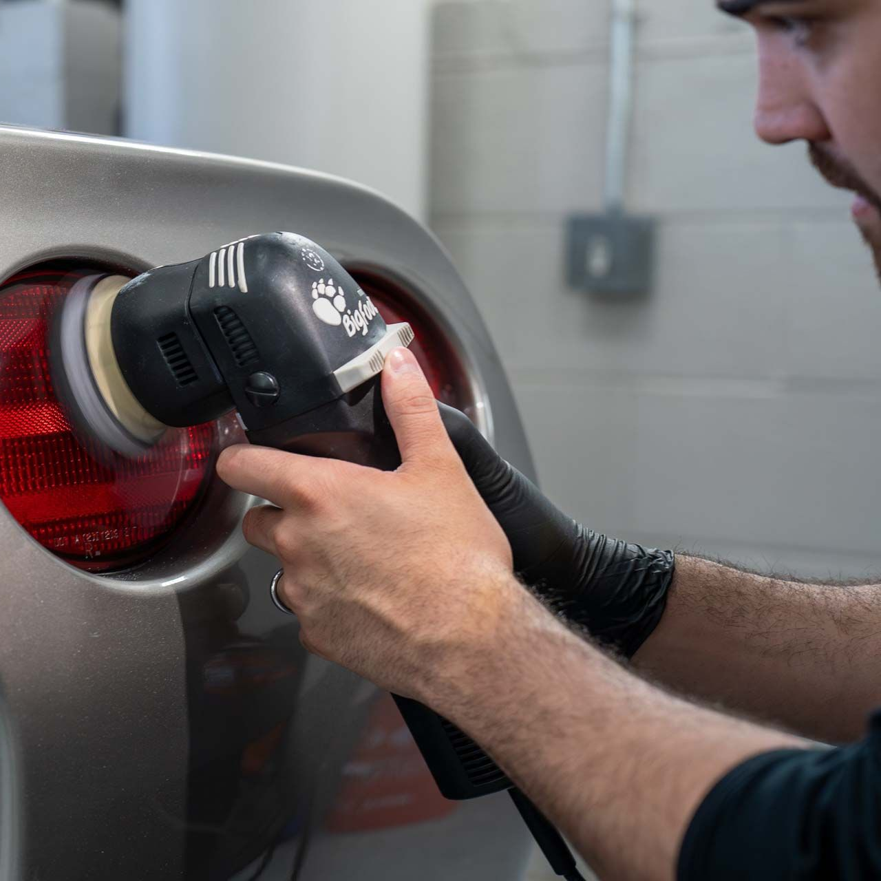 Person polishing a silver car’s red taillight with a handheld buffer in a workshop