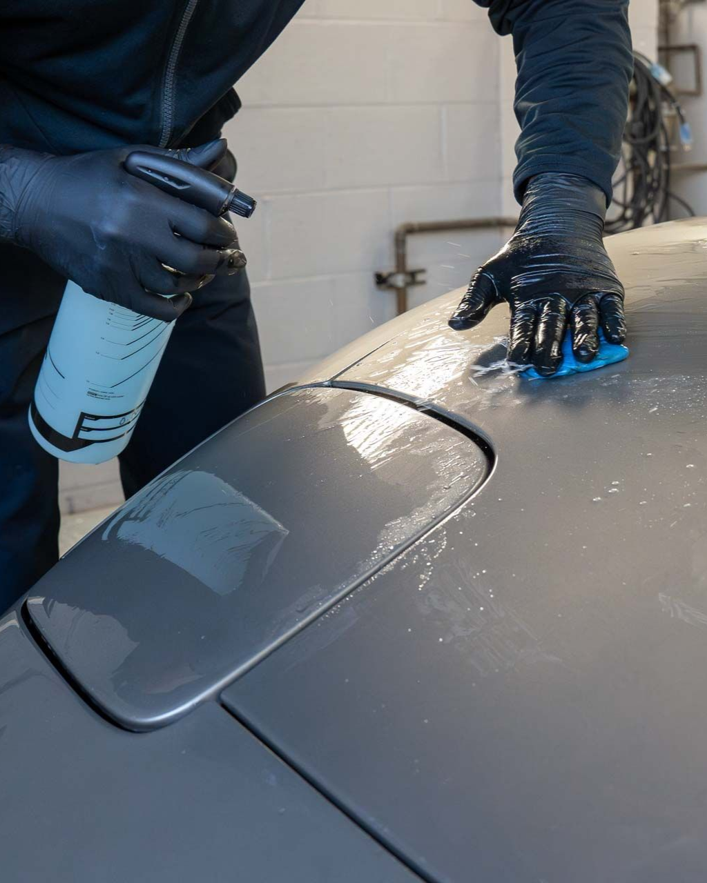 Gloved person polishing a gray car with a spray bottle and cloth in a garage