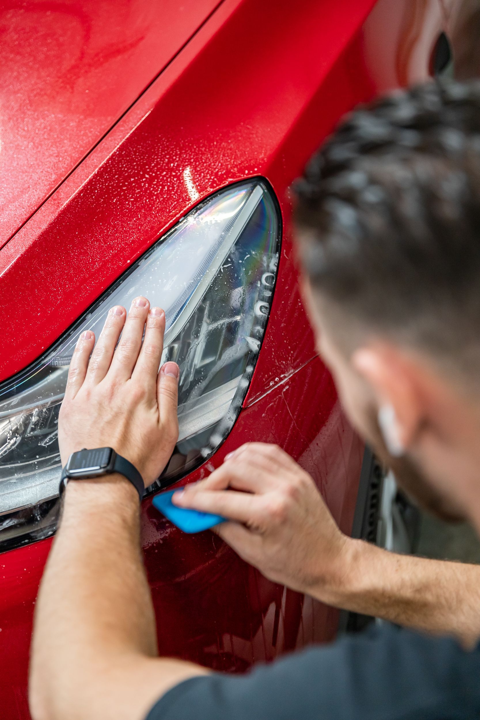 Person smoothing red car paint near a headlight with a blue tool