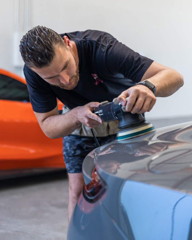 Detailing car paint with a polisher in a garage, man leaning over a blue vehicle.