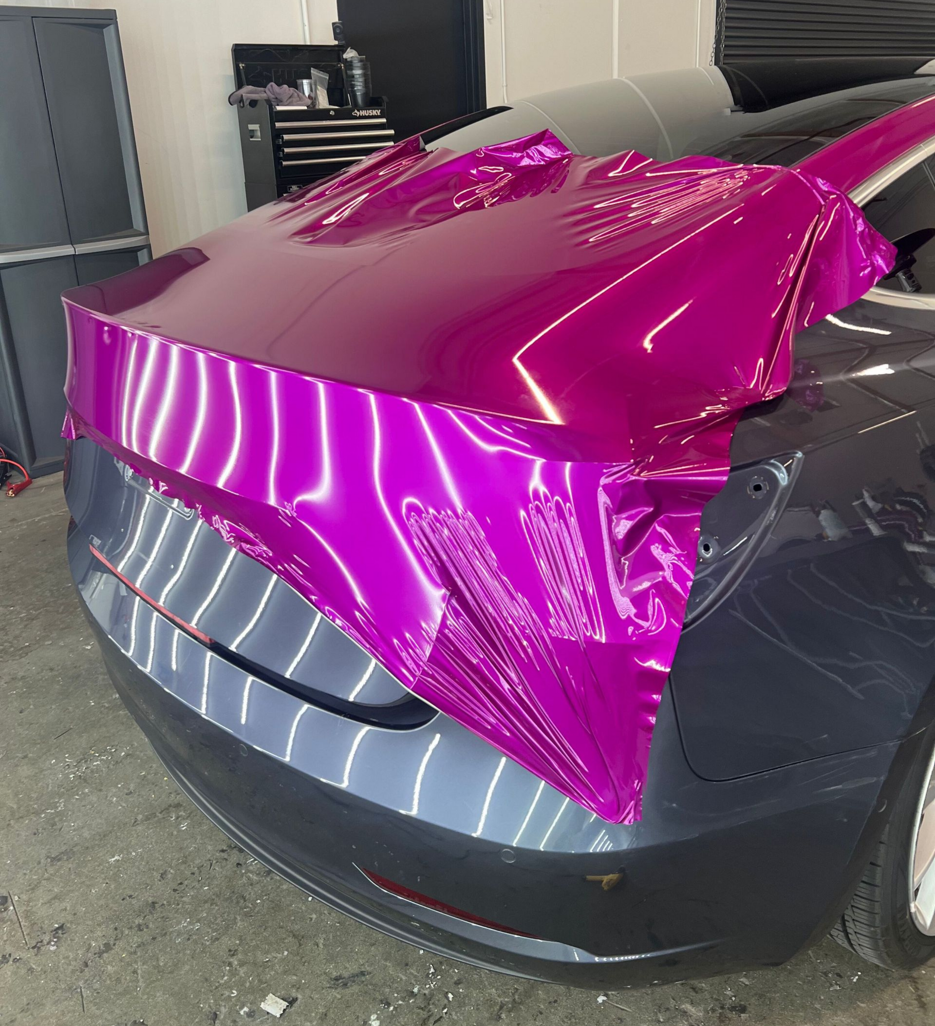 Magenta vinyl wrap being applied to the rear of a dark gray car in a garage