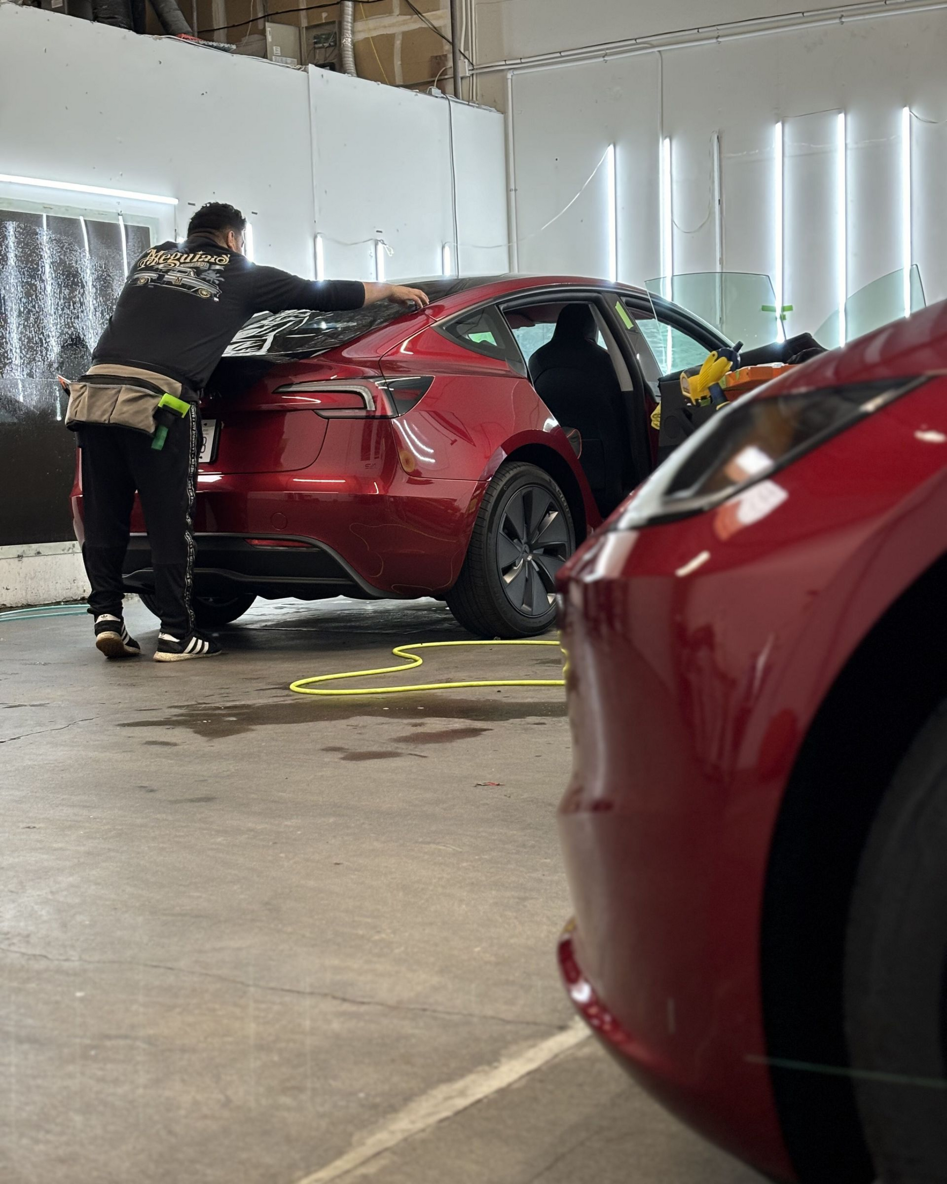 Red car being inspected in a garage, with a person leaning over the rear and another red car in the foreground.