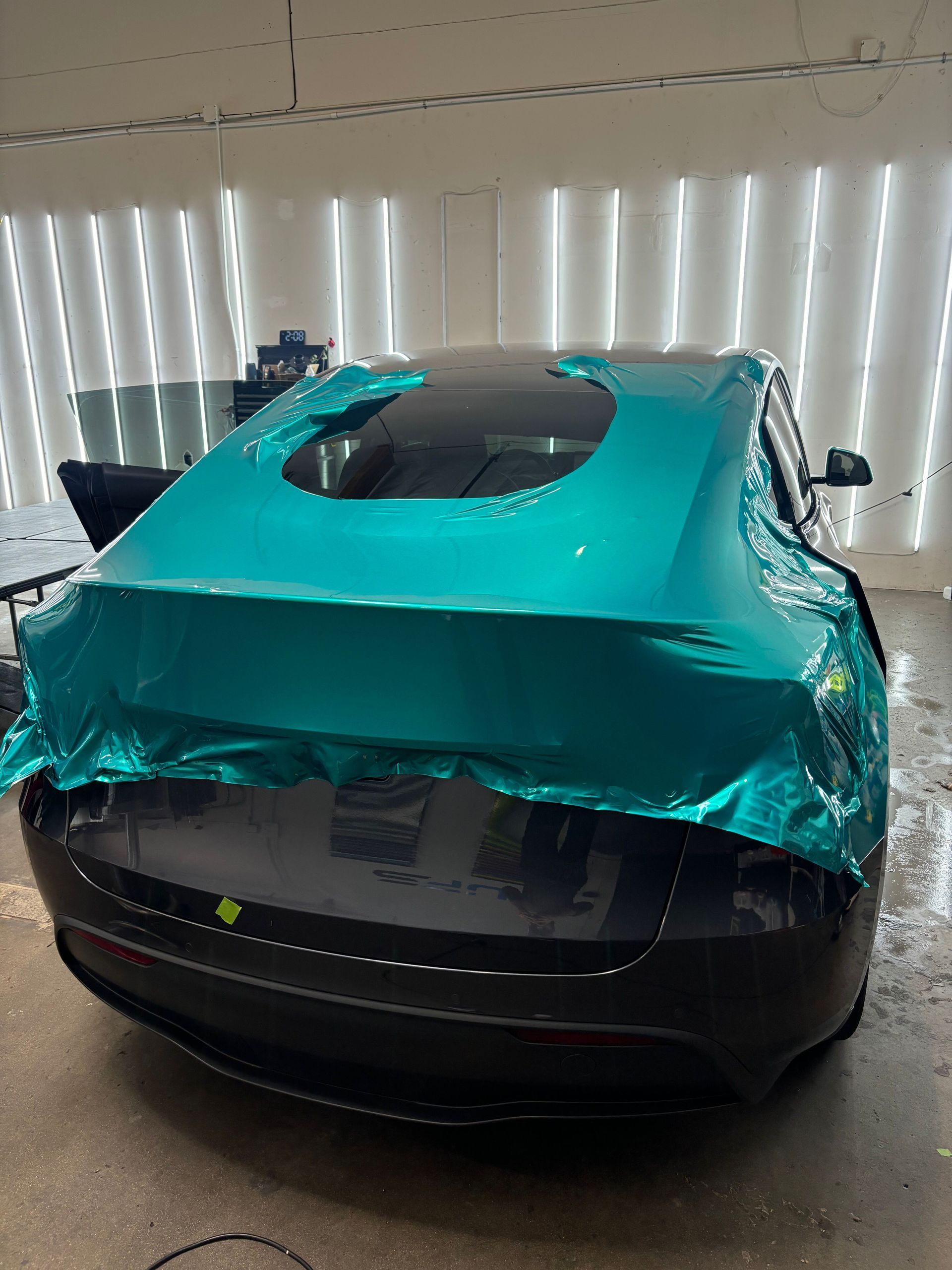 Black car wrapped in teal protective film inside a garage with vertical blinds