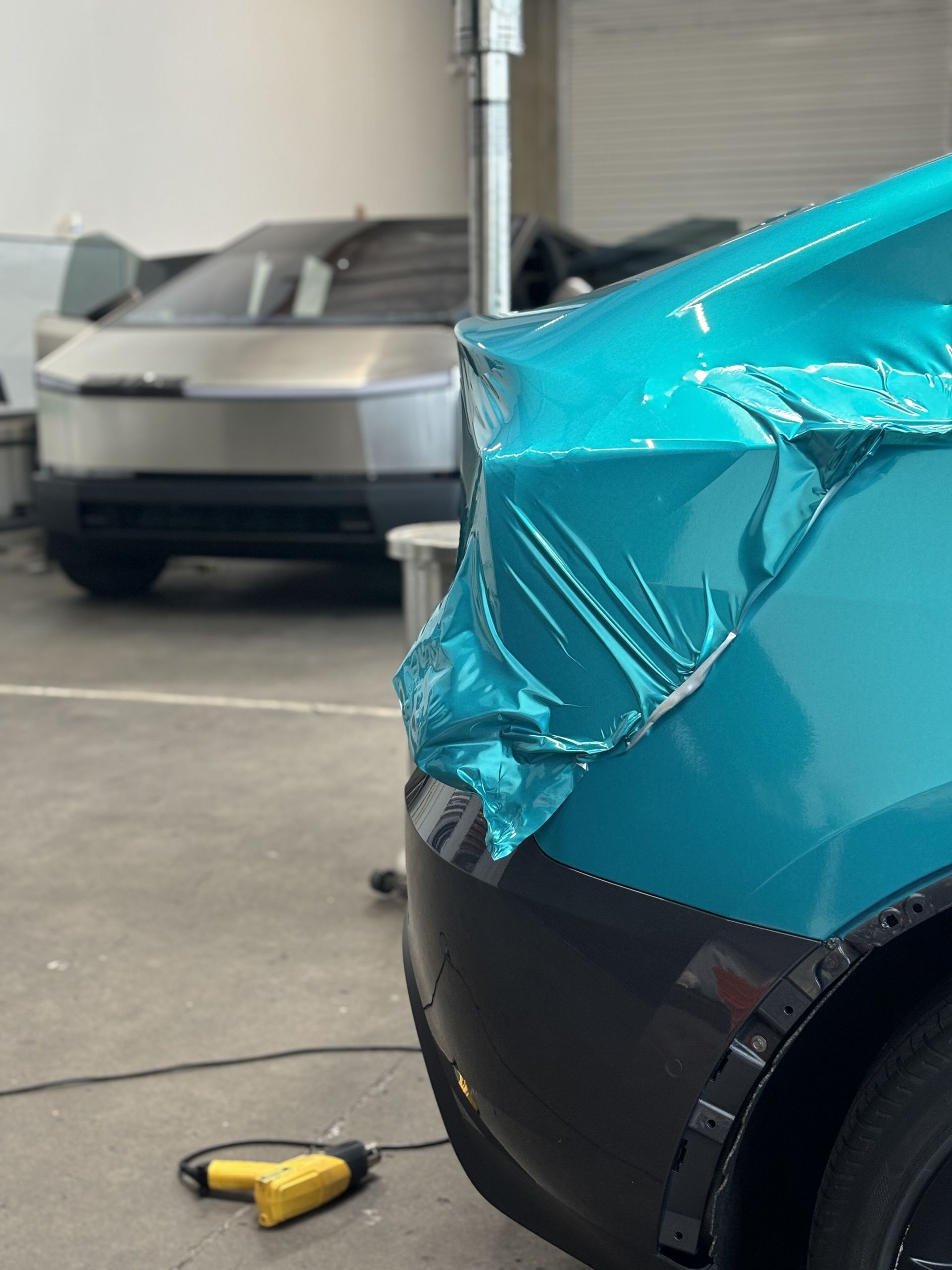 Crushed turquoise car front corner in a garage, with a silver car and yellow tool in the background.