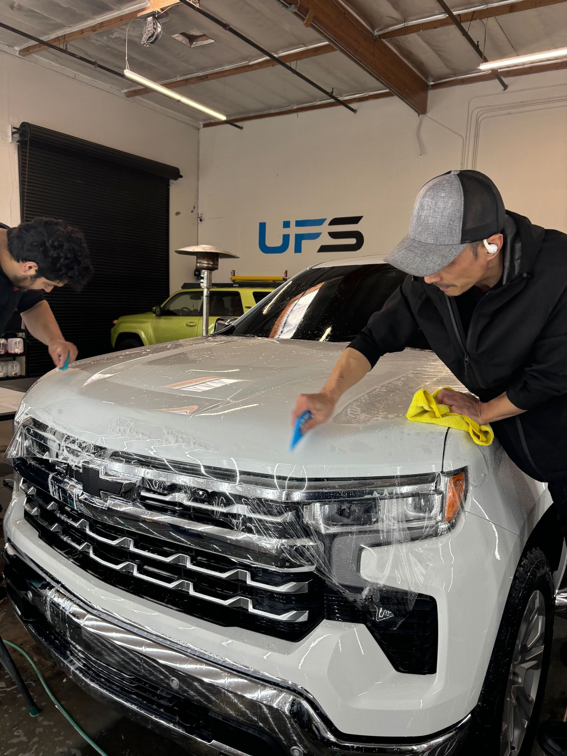 Two people detailing a white SUV in a garage, wiping the hood and windshield.