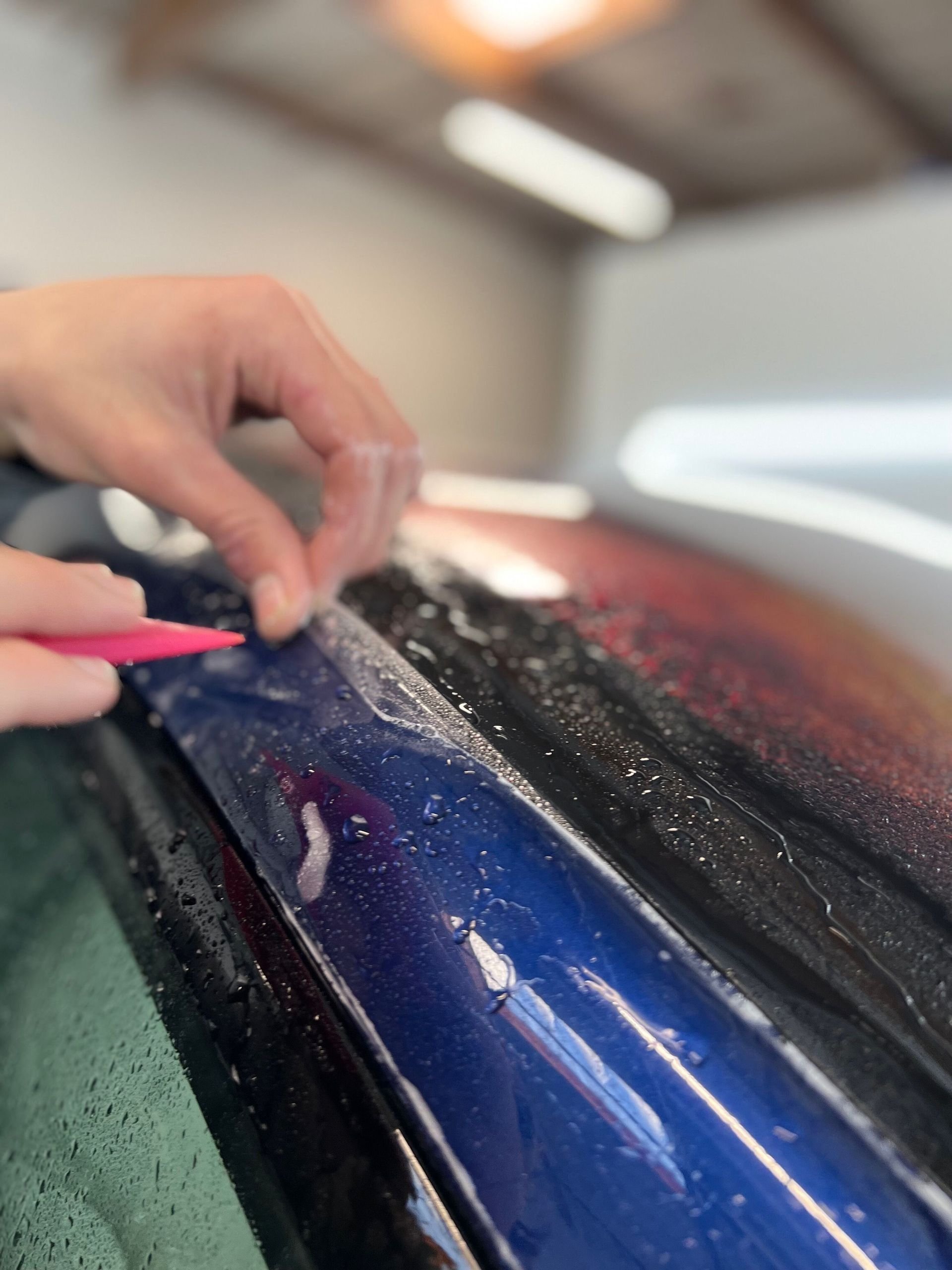 Hands polishing a blue car surface with a cloth and pink tool in a garage