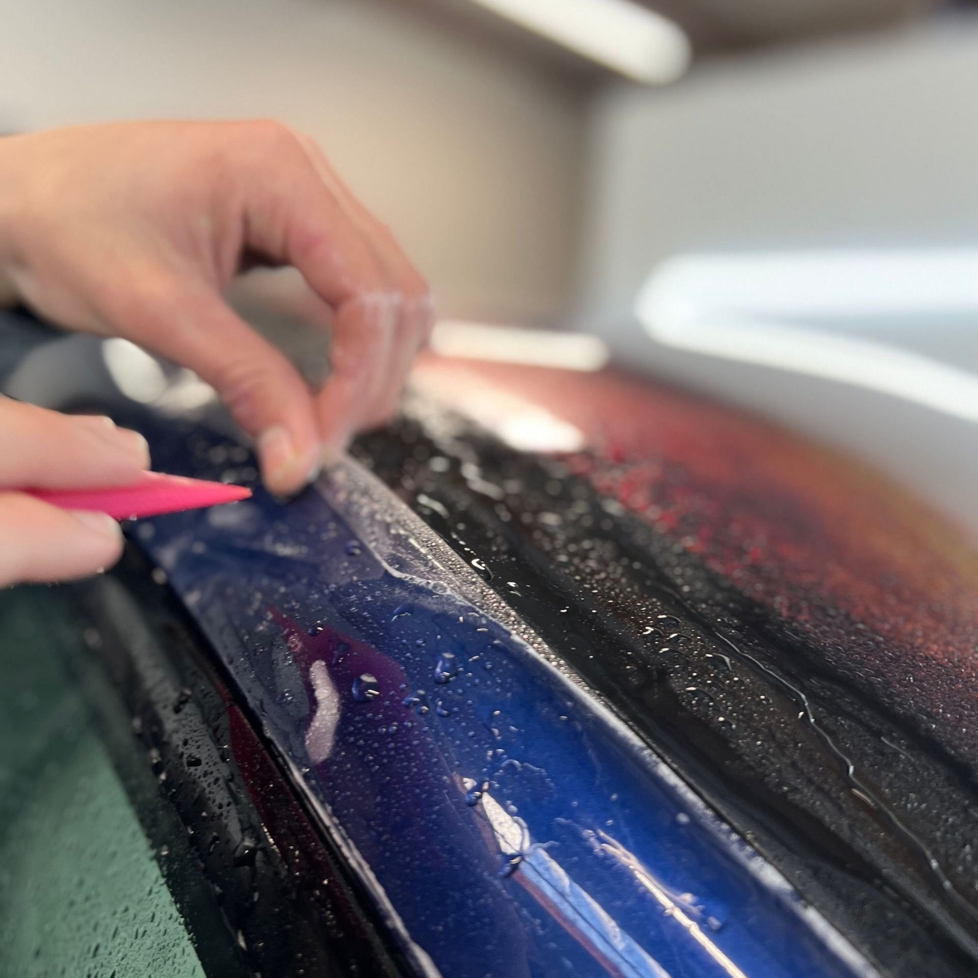 Hands wiping water off a blue car hood with a pink cloth in a garage