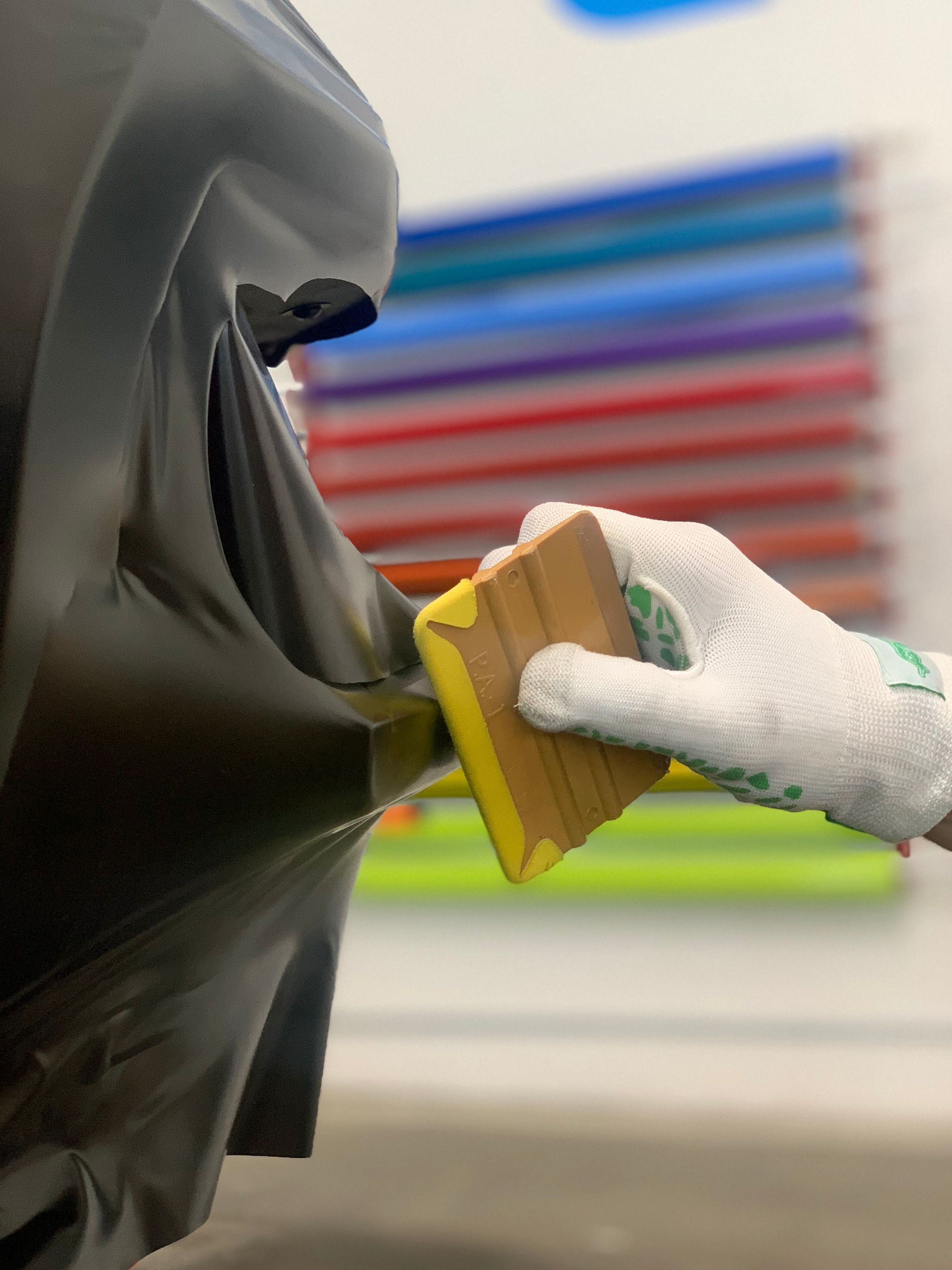 Gloved hand sanding a black car panel with a yellow sanding block in a workshop