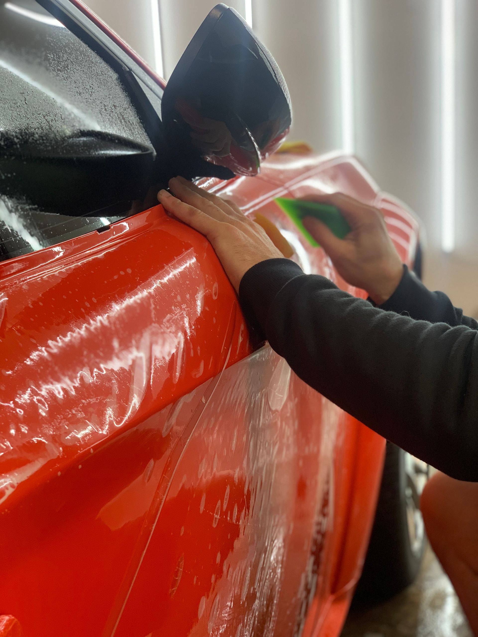 Hands smoothing red vinyl on a car, with black and pink sections visible.
